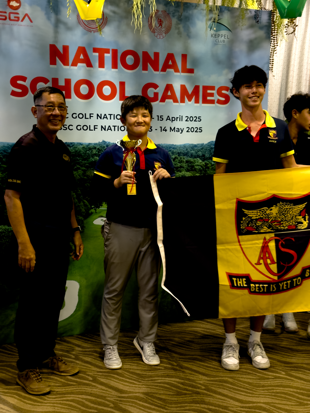 Three people pose at National School Games golf event, one youth holding a trophy and another displaying a yellow ACS school flag.