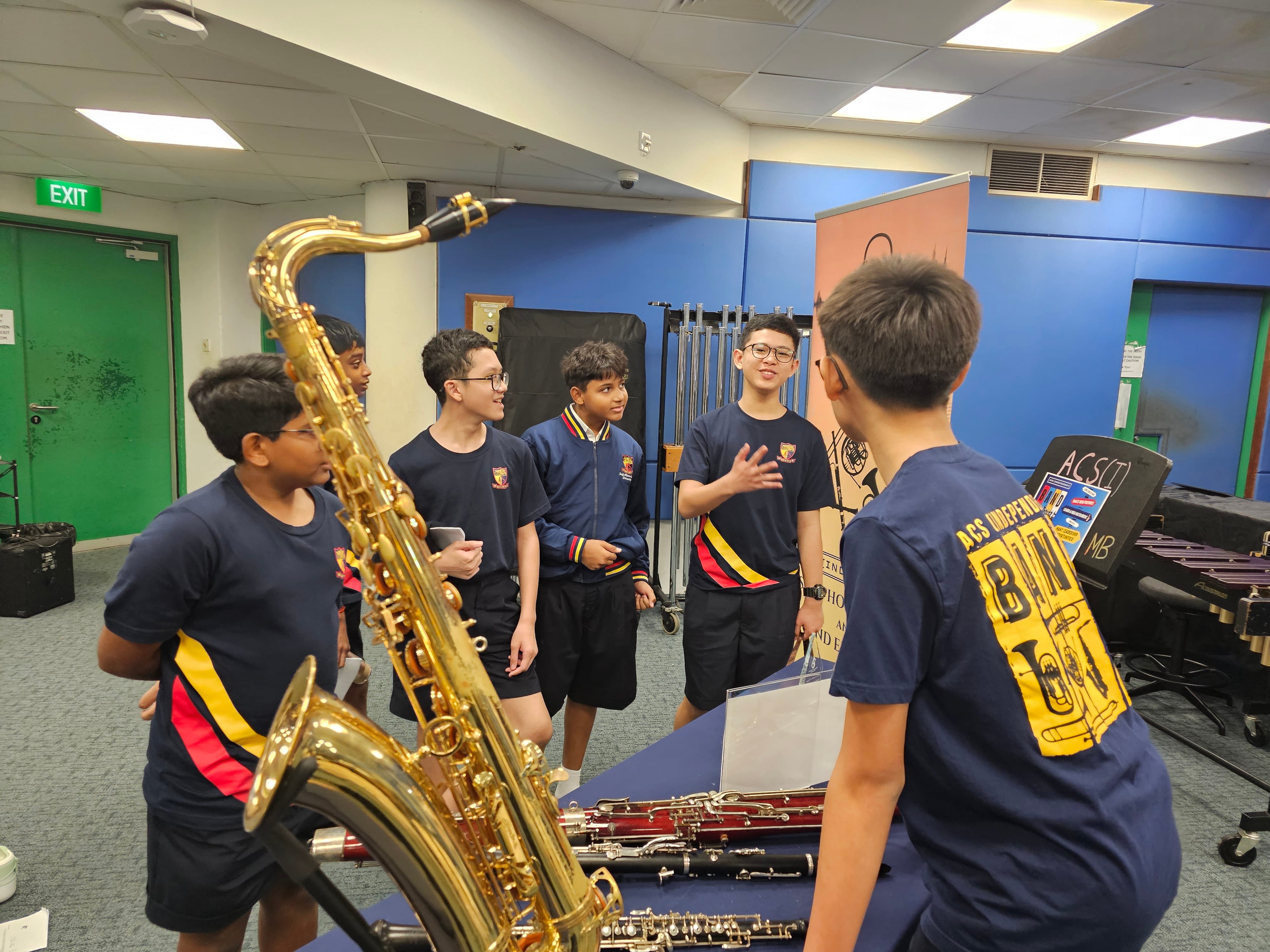 Six boys in navy school uniforms stand in a music room near a large saxophone and clarinets on a table.