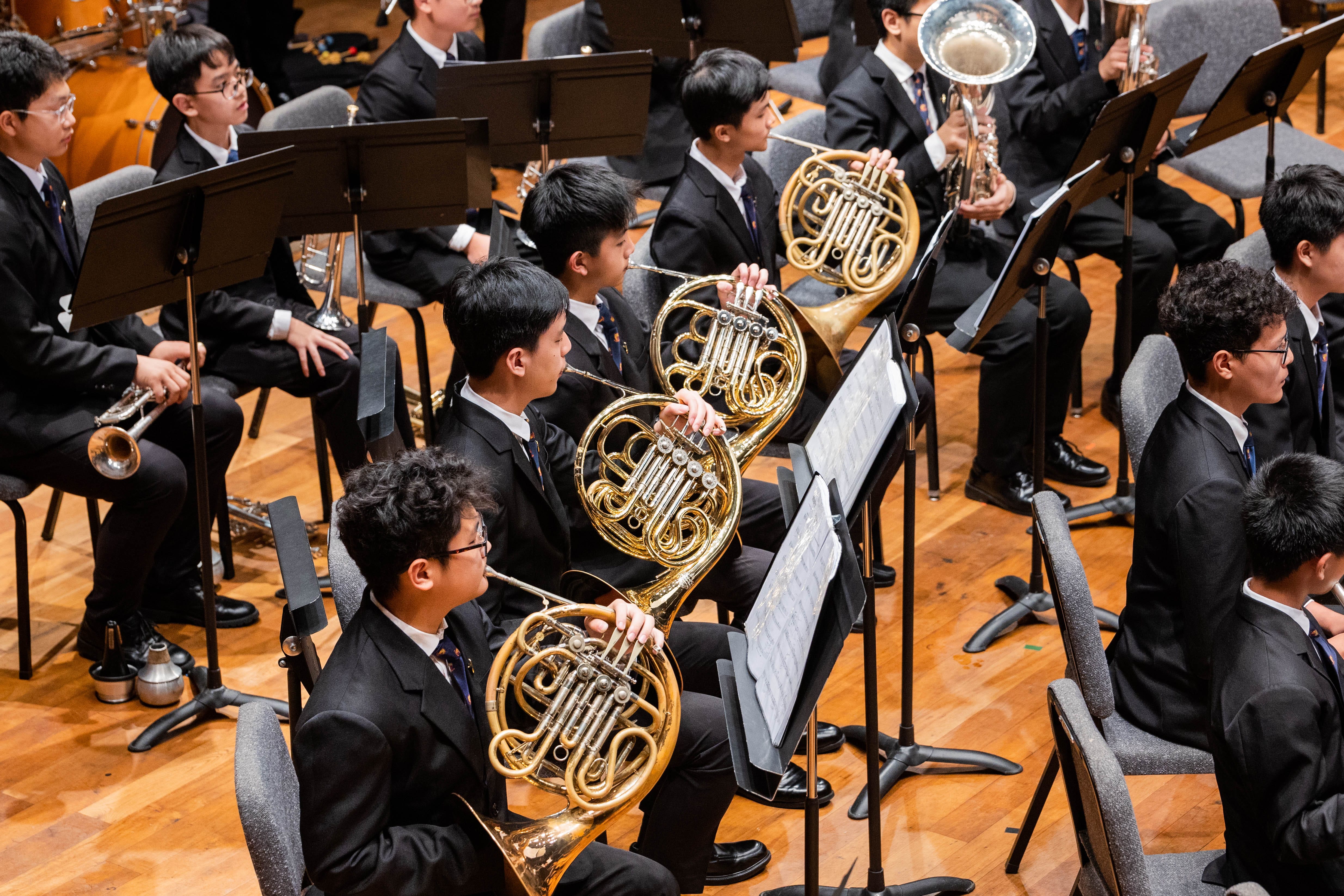 Young musicians in black suits play French horns and trumpets in a seated orchestra on a wooden stage.