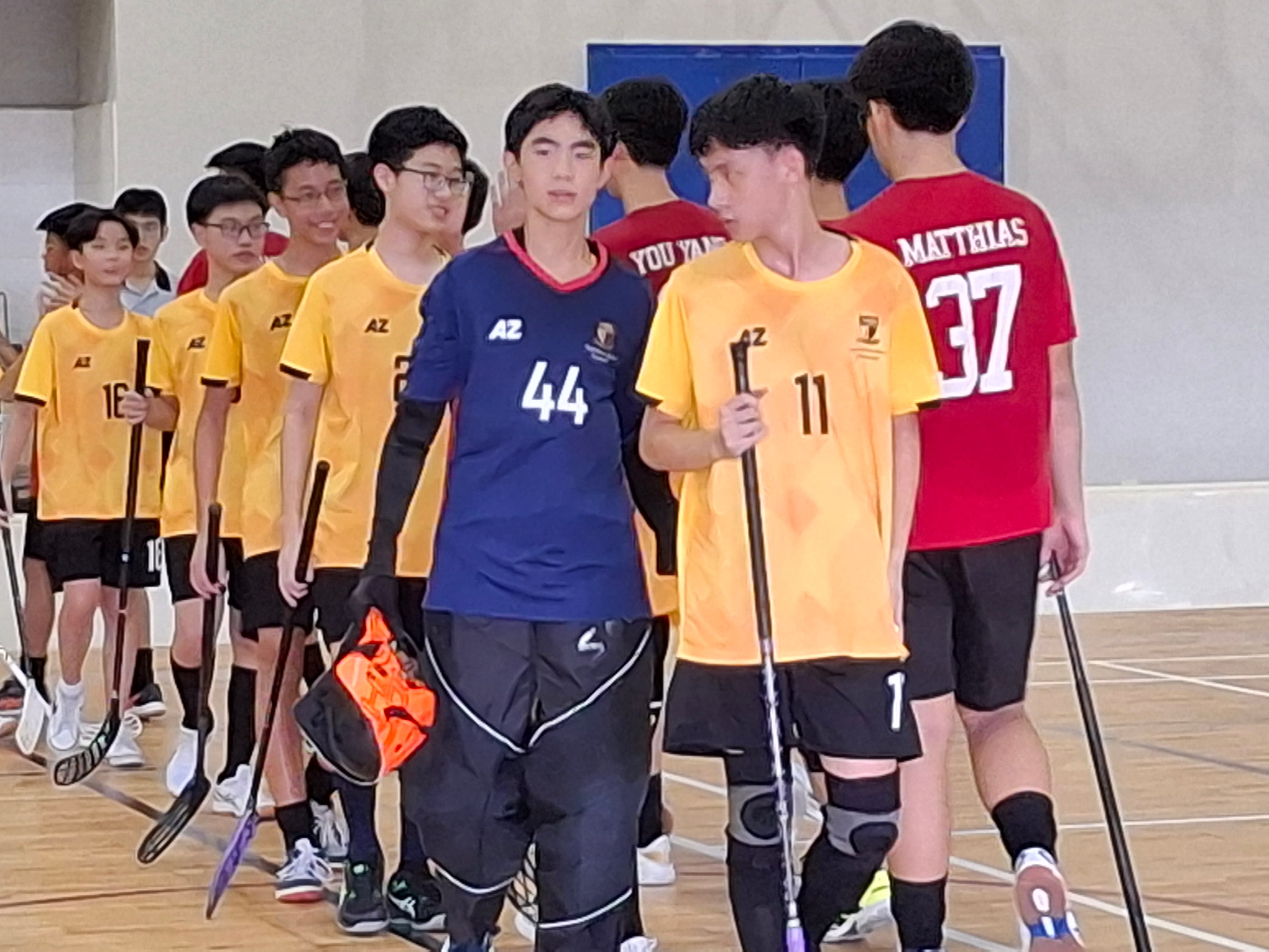 Teen boys in yellow and blue numbered jerseys holding floorball sticks walk together in an indoor gymnasium.