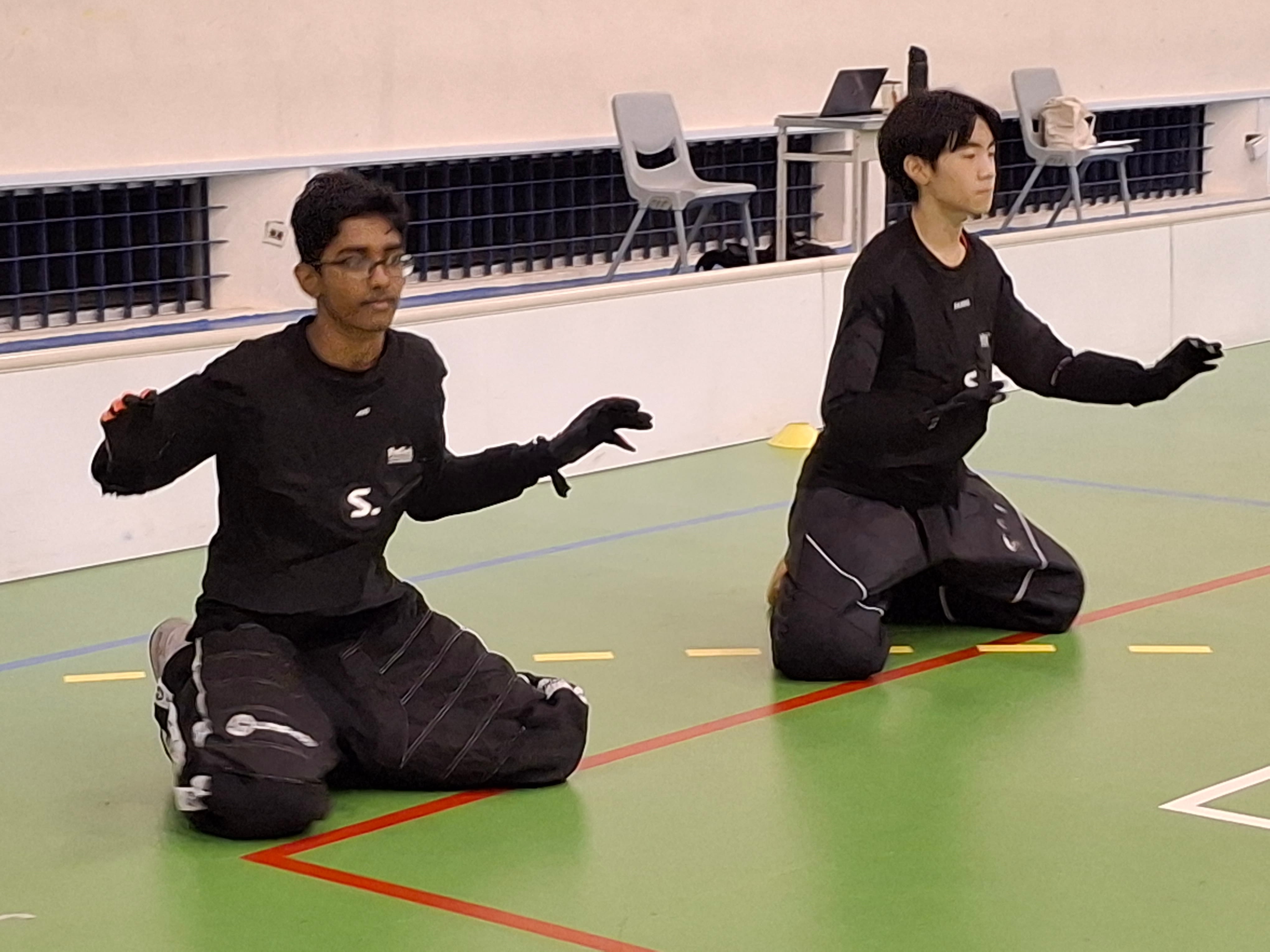 Two young men in black outfits kneel on a green gym floor with arms outstretched in a synchronized pose.