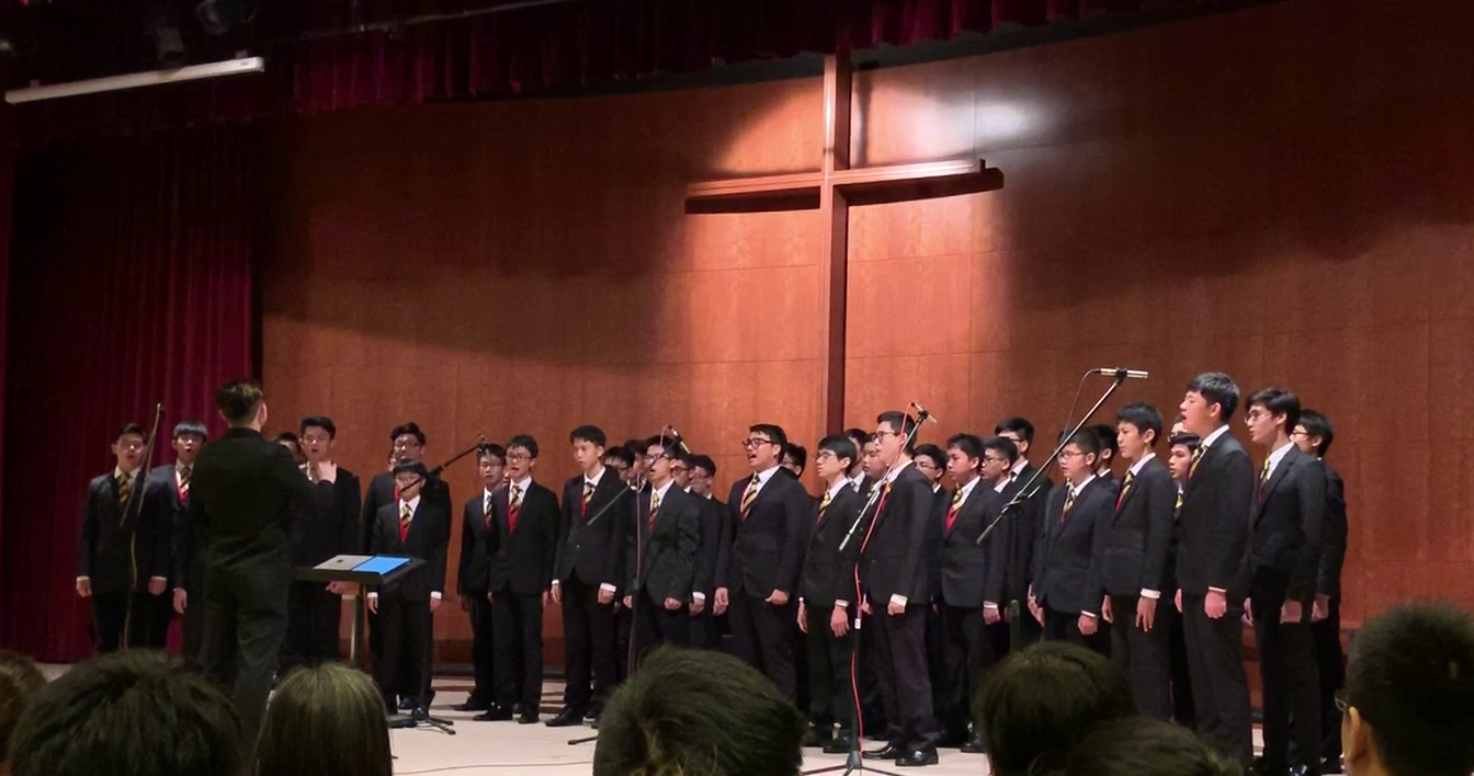 A boys choir in dark suits and striped ties performs on stage before a wooden cross, led by a conductor.