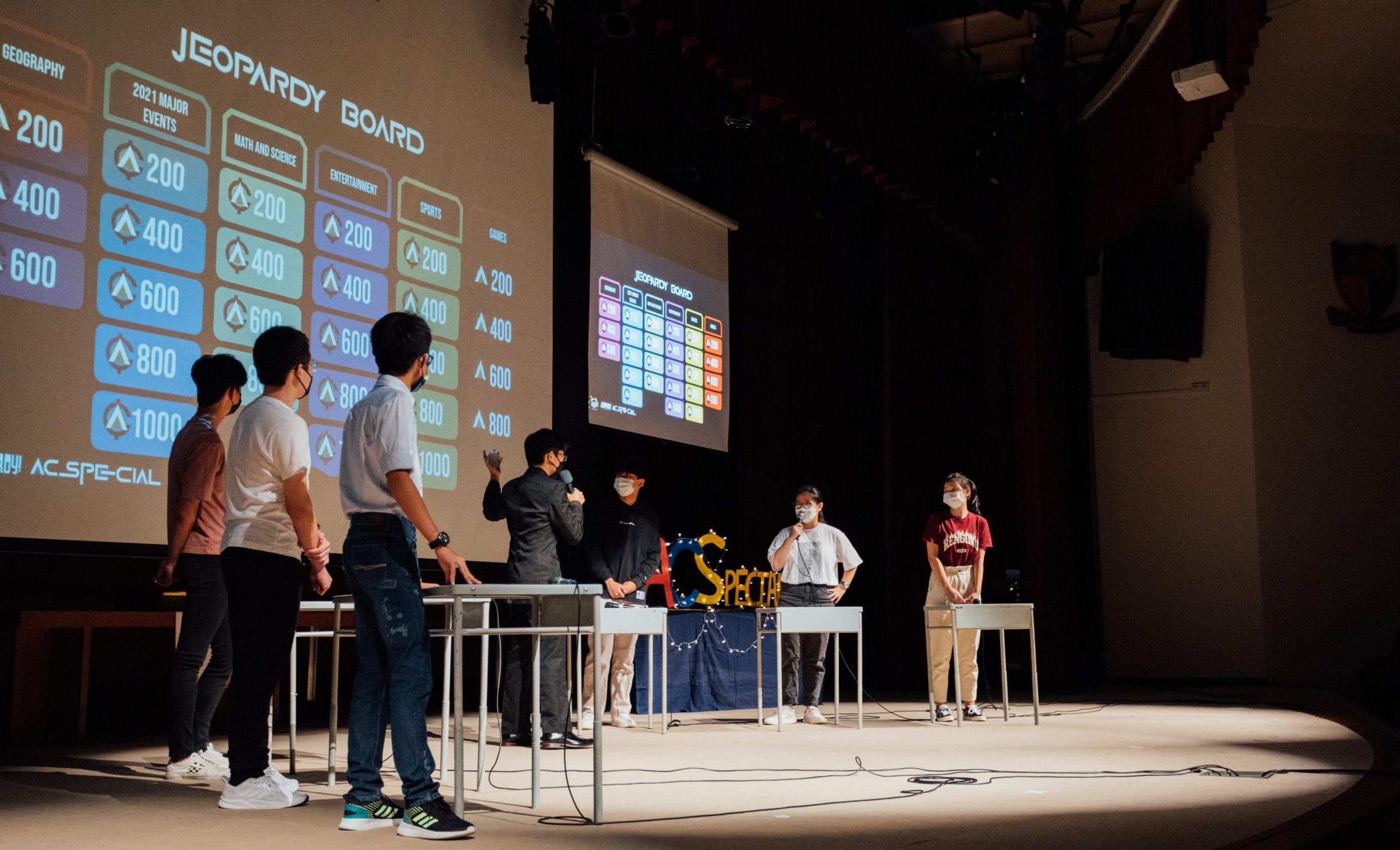 Students wearing masks stand on a stage playing Jeopardy, with a large Jeopardy Board projected on screens behind them.