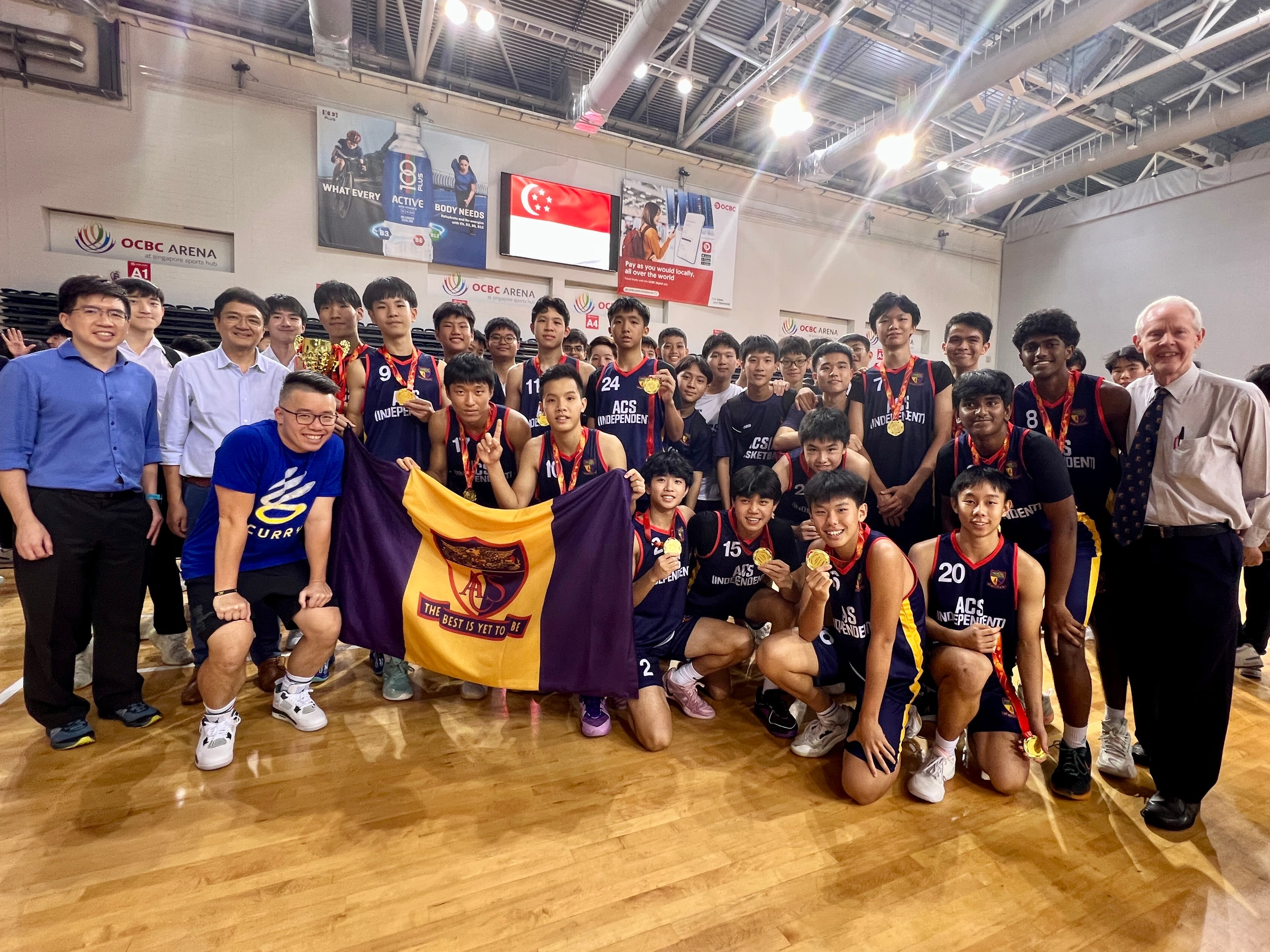 ACS Independent basketball team posing with medals and a school flag at OCBC Arena in Singapore.