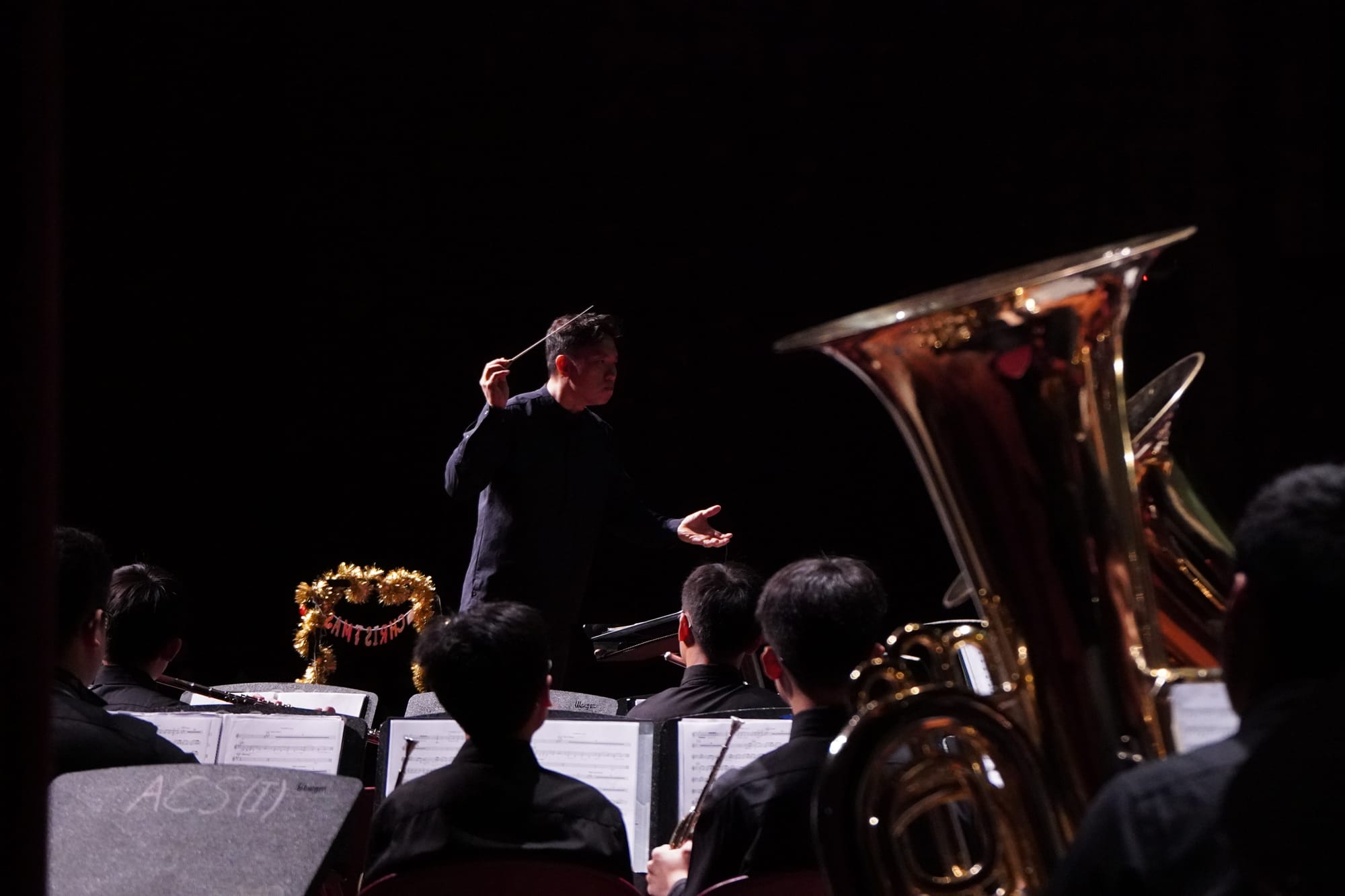 A conductor in black raises a baton over seated musicians with brass instruments and sheet music on stands.