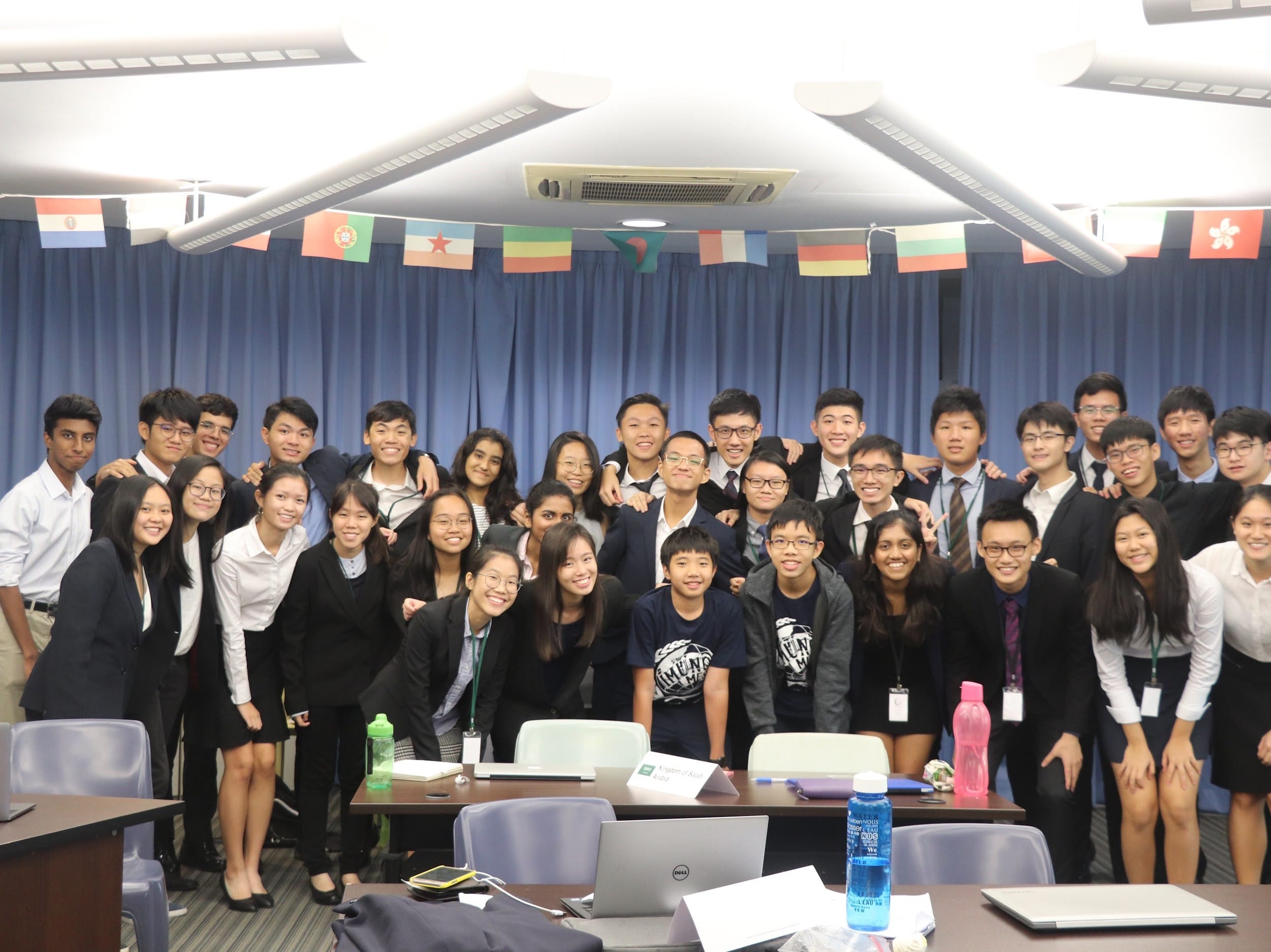 A large group of ACS(I) Young Diplomats Society members in formal black and white attire poses for a group photo in a conference venue decorated with international flags.