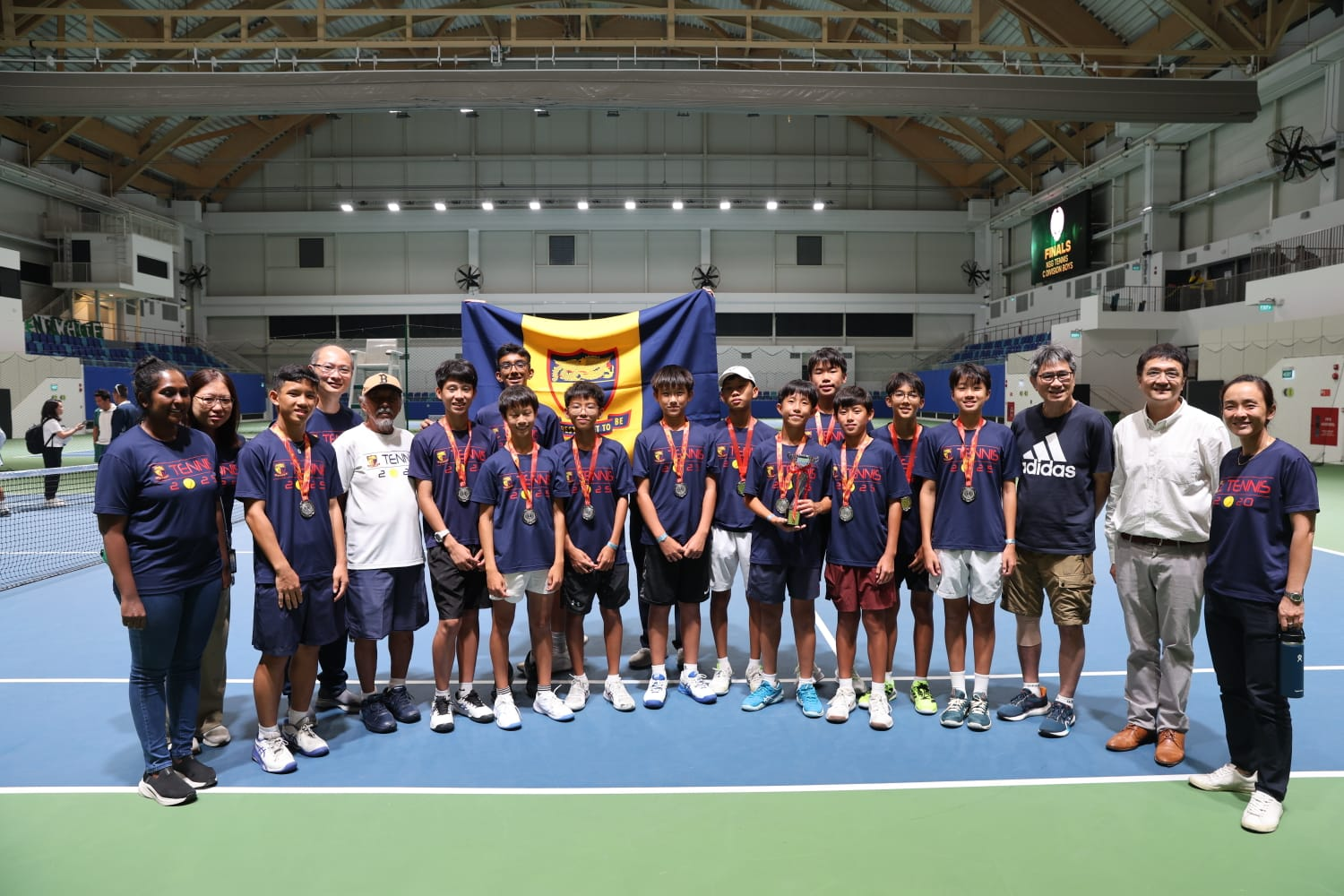 An indoor photo of the tennis team with the school flag in the background.