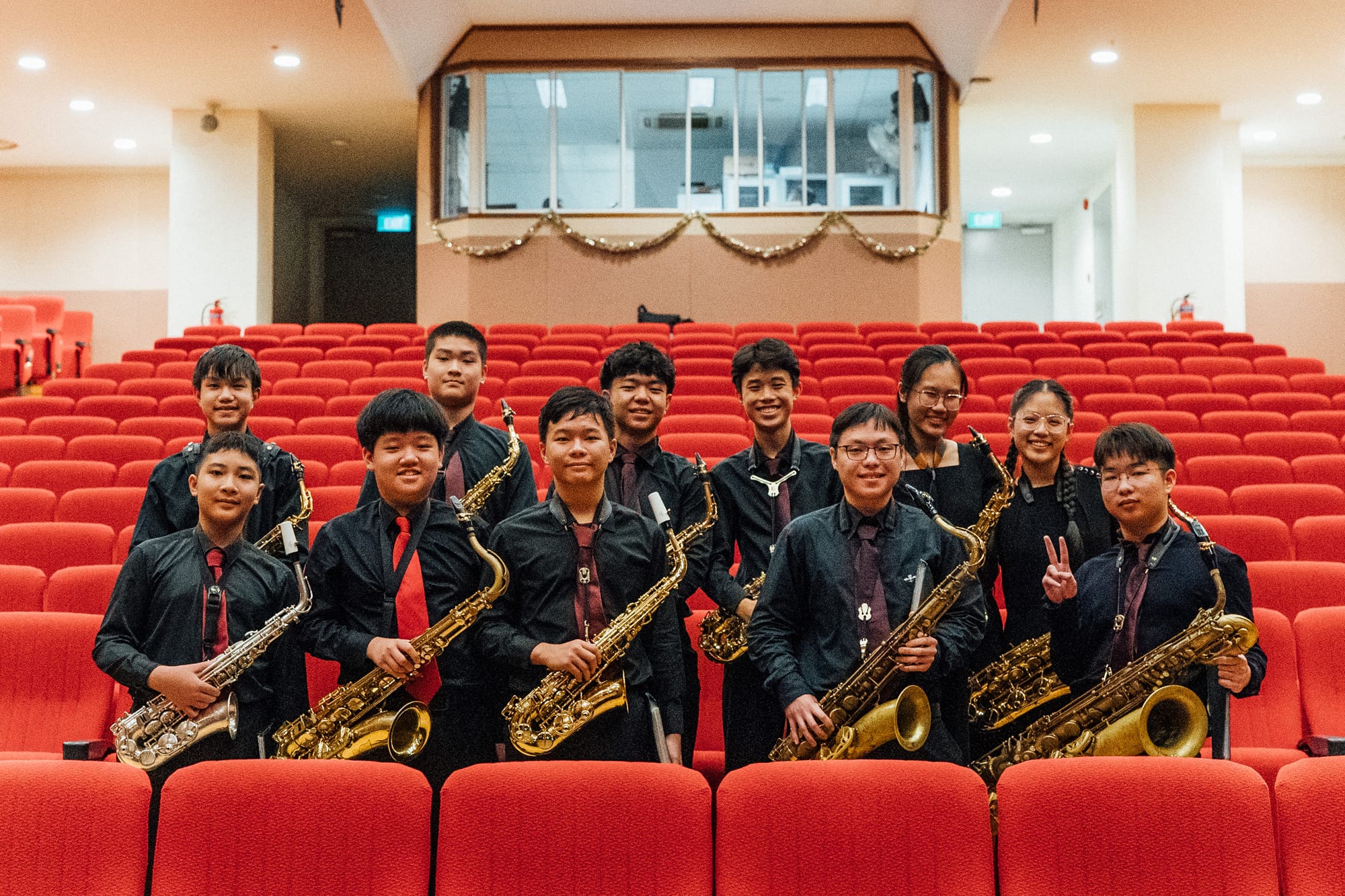 A group of young musicians in black shirts holding saxophones, posing in rows of red auditorium seats.