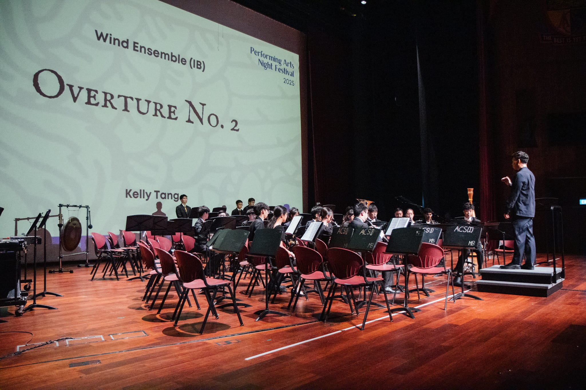 A wind ensemble performs Overture No. 2 on stage at a 2025 Performing Arts Night Festival, led by a conductor.