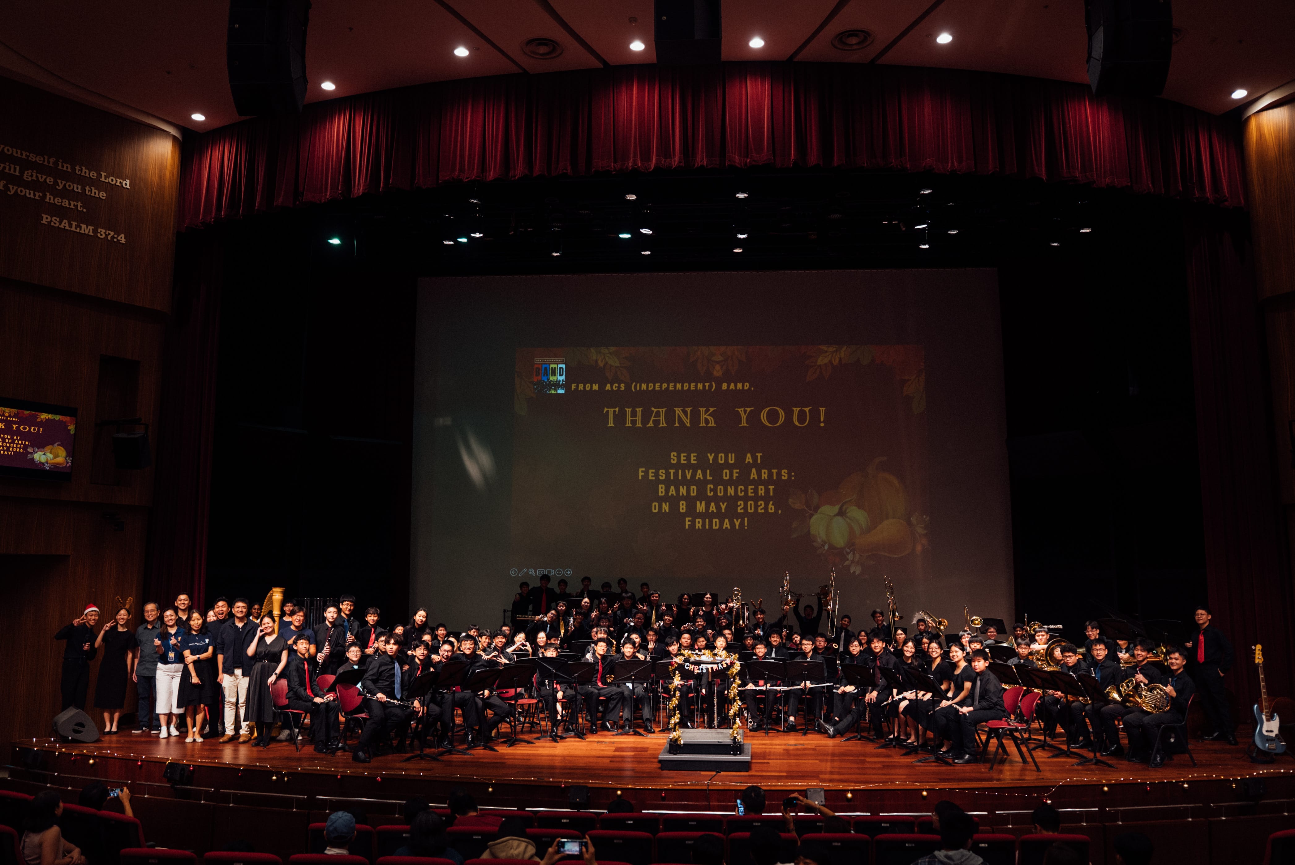 Large school band posed on a concert hall stage with a Thank You screen, red curtains, and audience in front.