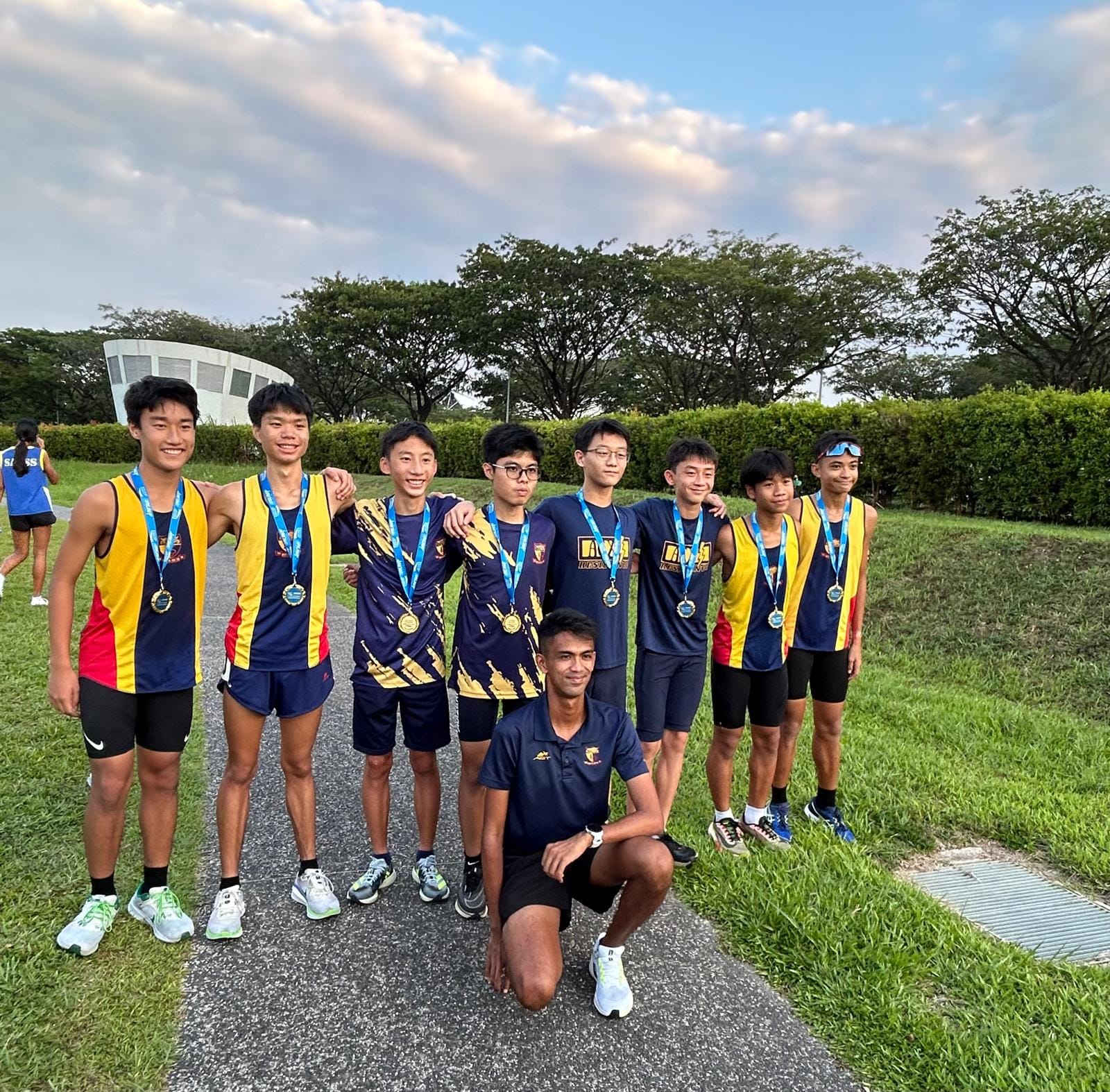 Eight young male runners wearing medals pose with a coach kneeling in front, outdoors near trees.