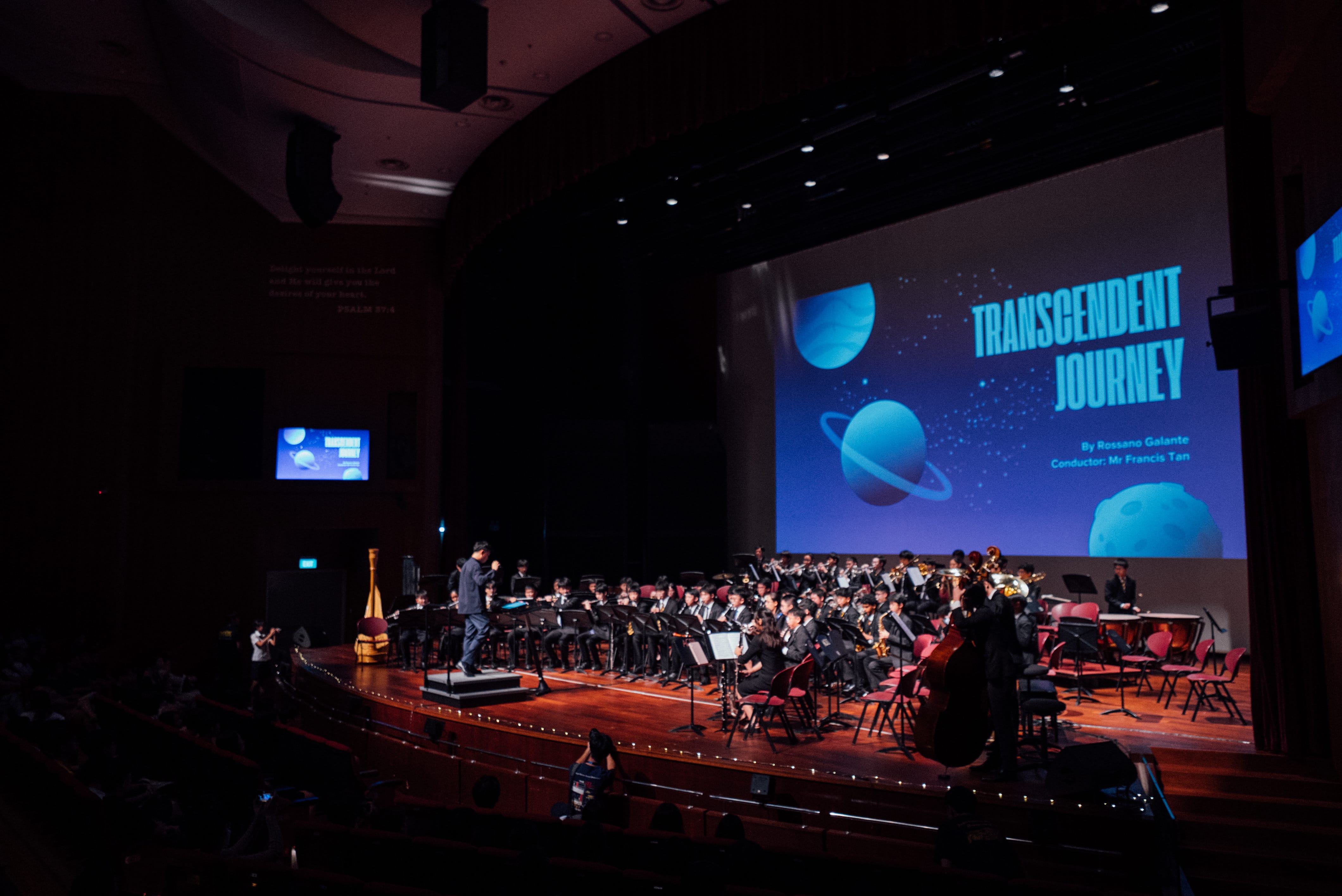 A conductor leads a large student band on a dark stage with a screen reading "Transcendent Journey" behind them.