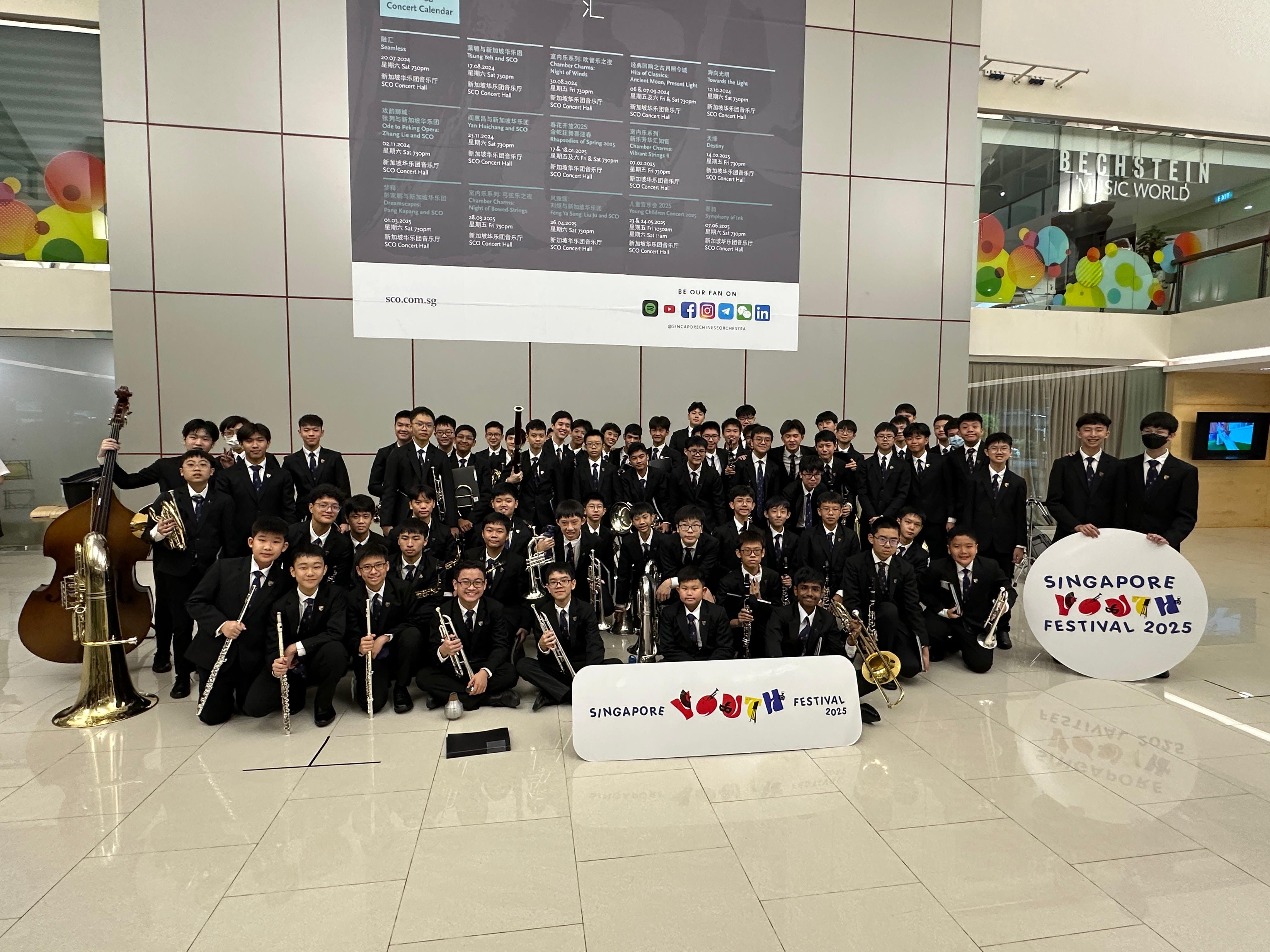Large youth orchestra in black suits posing with instruments and Singapore Youth Festival 2025 signs indoors.
