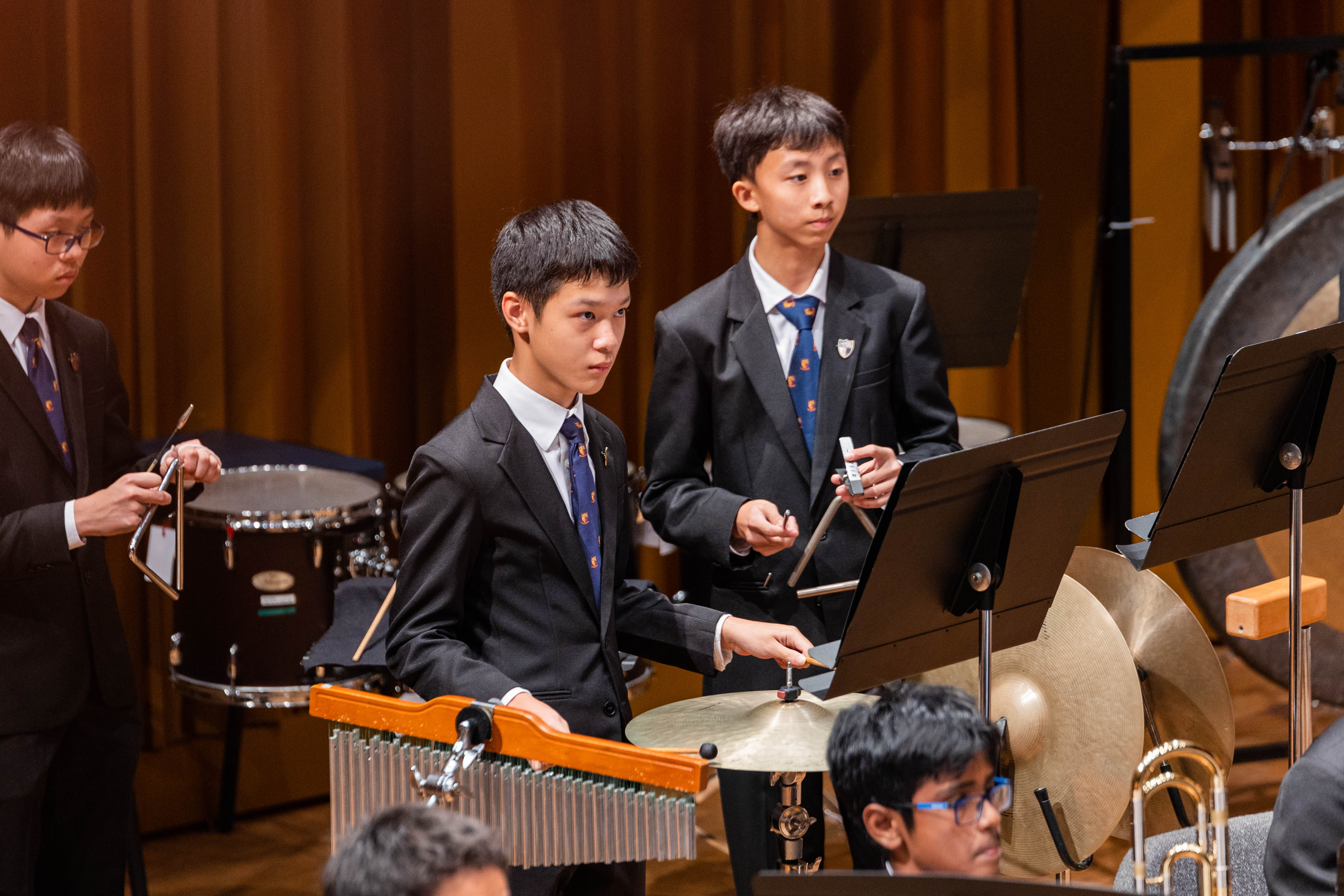 Three young male students in suits and ties perform percussion instruments including chimes, triangle, and cymbals on stage.