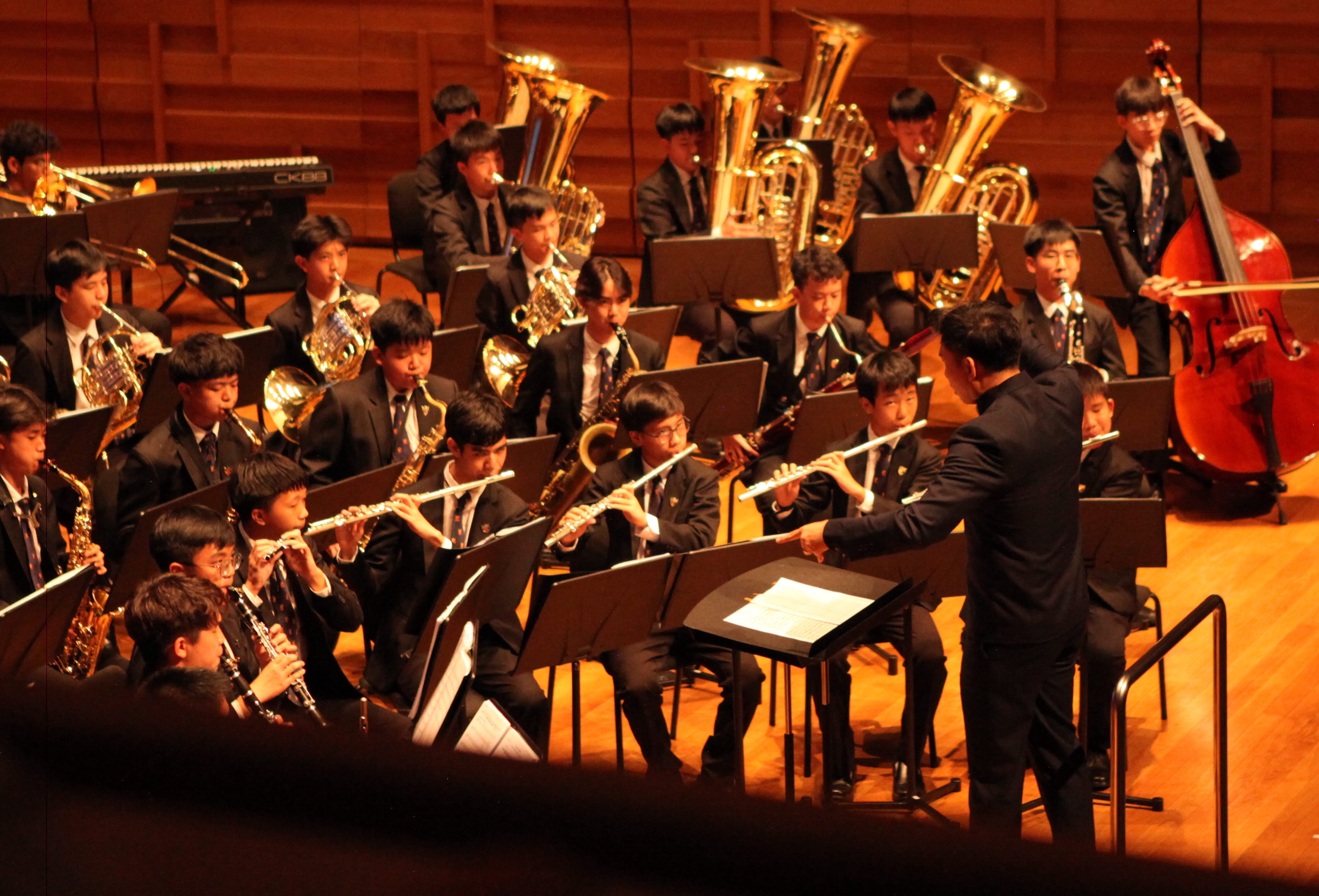 A conductor leads a youth wind band playing flutes, brass, and woodwinds on a warmly lit concert stage.
