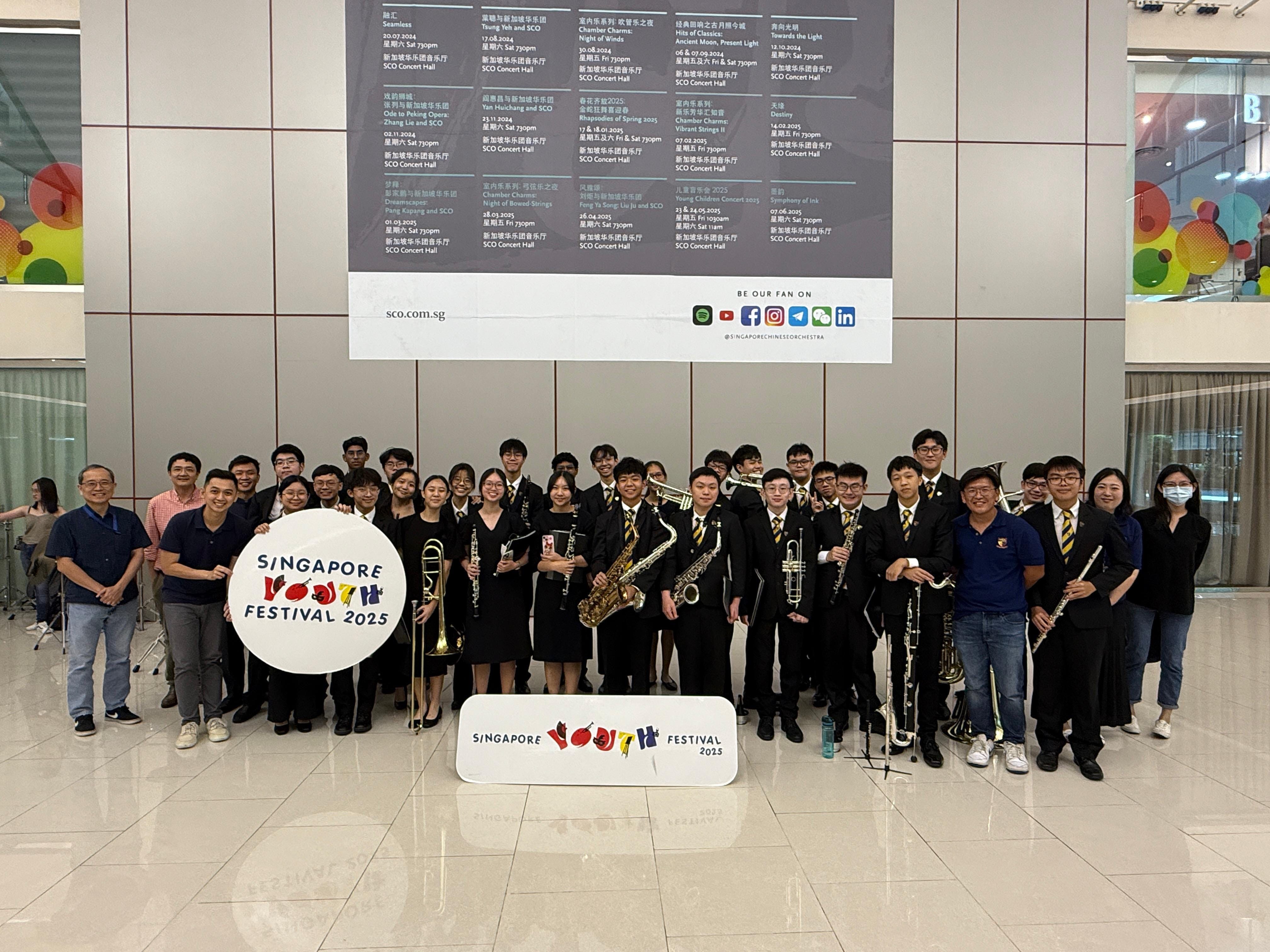A large group of young musicians in black uniforms pose with instruments at Singapore Youth Festival 2025.