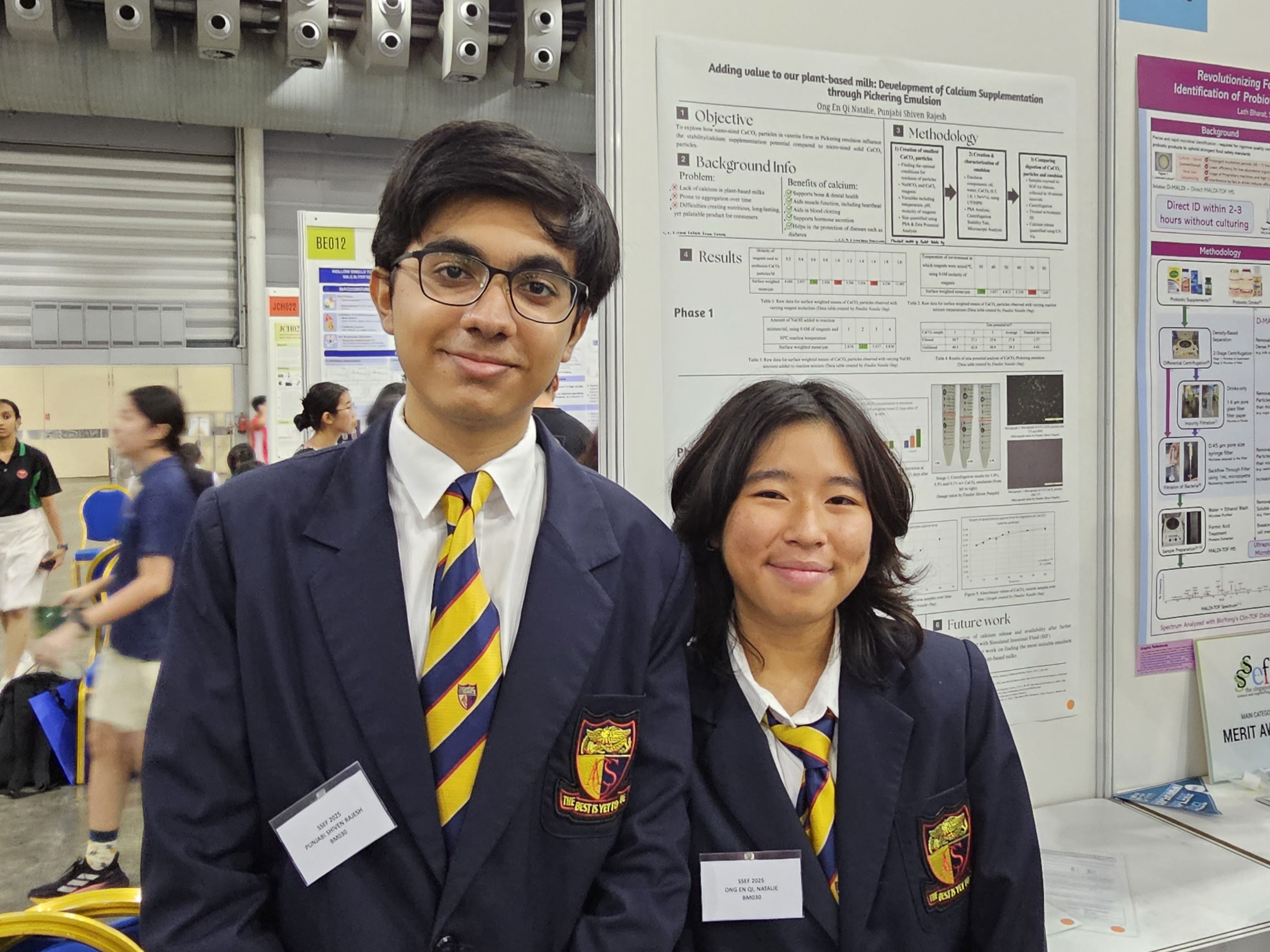 Two students in navy blue blazers over their school uniforms stand smiling in front of a science fair project poster at an indoor event.