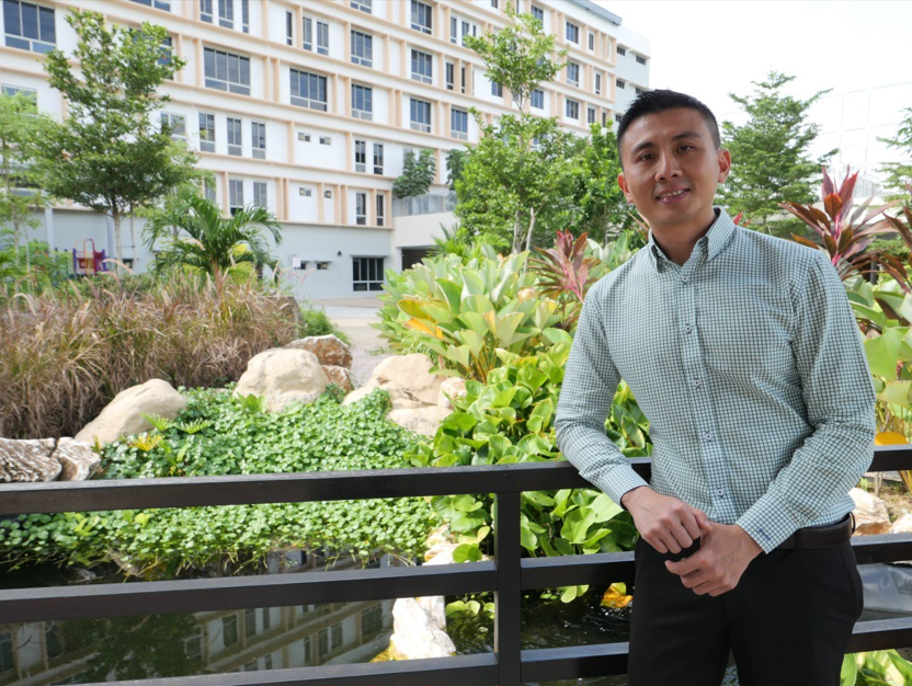 Man in a light checkered shirt stands by a railing in front of a lush garden with rocks and a multi-story building.