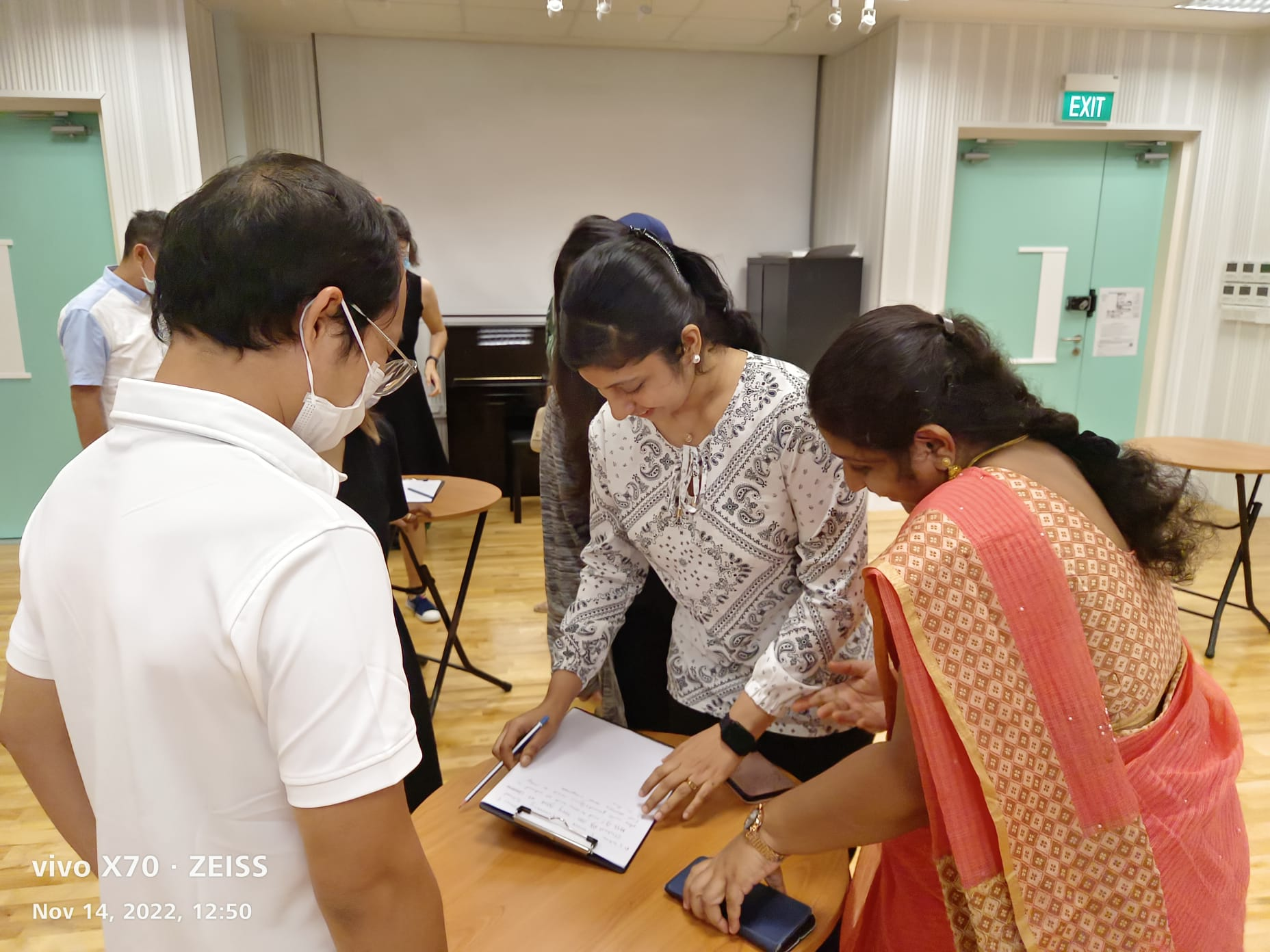 Three parent volunteers huddle around a clipboard on a table in a room, with one woman writing on a paper.