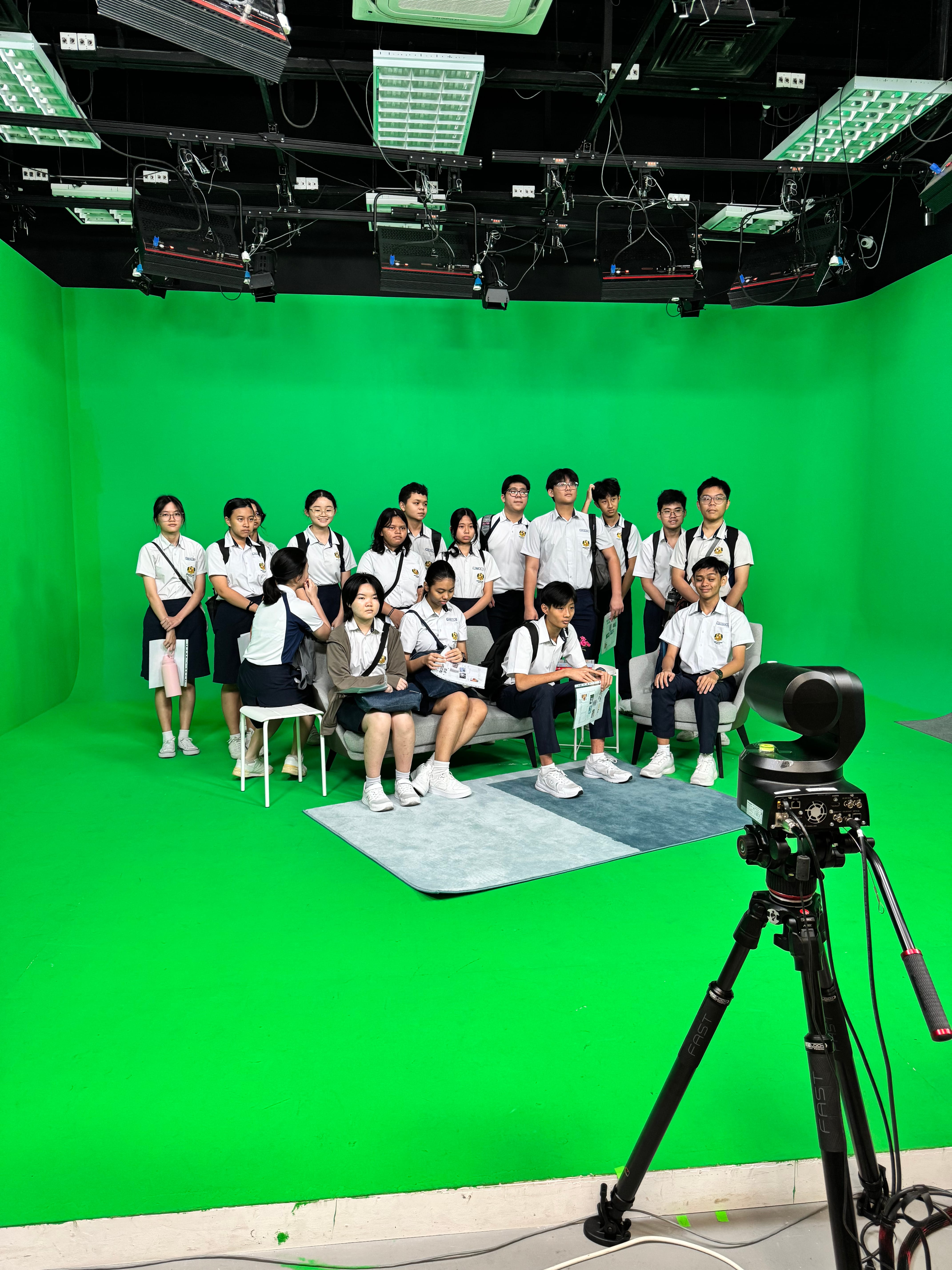 Group of students in uniforms on chairs in a green-screen studio, with ceiling lights and a camera tripod.