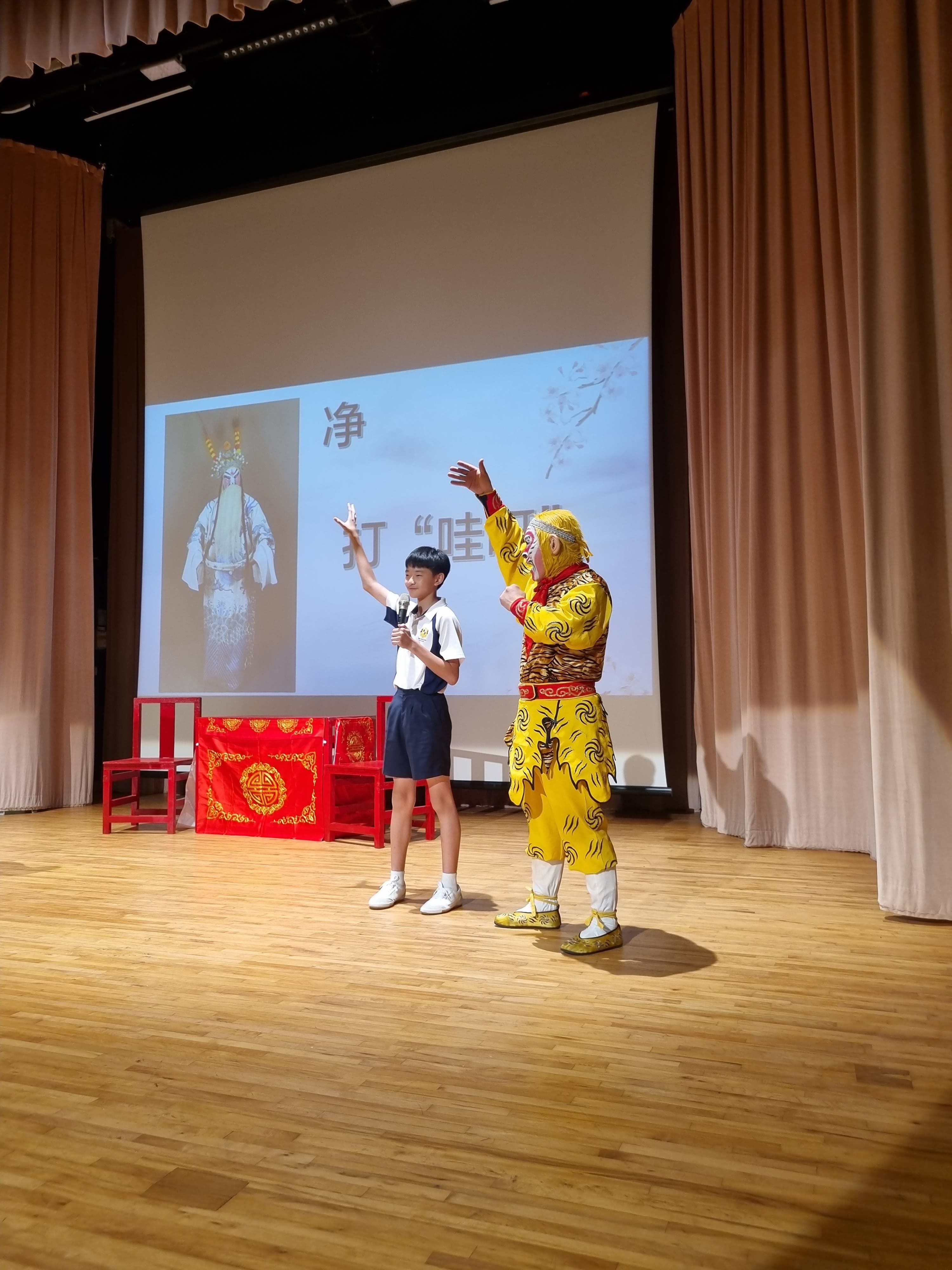 Two performers on a wooden stage, one in yellow costume and one in school uniform, arms raised by screen.
