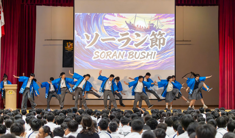 Students in blue robes perform Soran Bushi dance on stage before a large seated audience, with a projected screen behind them.