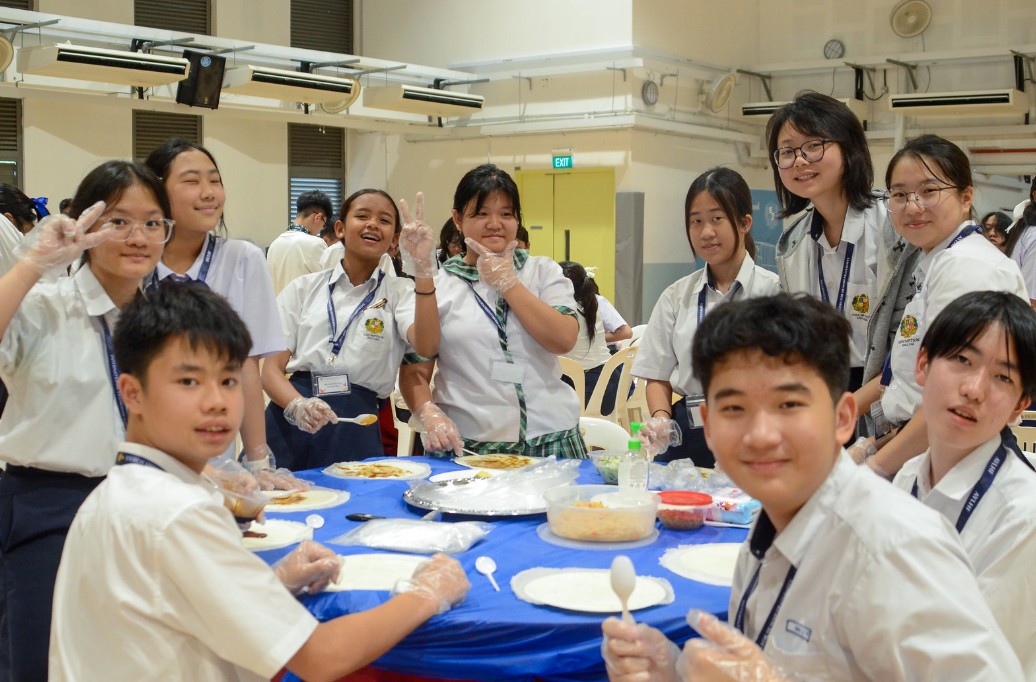 Students in school uniforms wearing plastic gloves gather around a blue-covered table with food during a cooking activity.