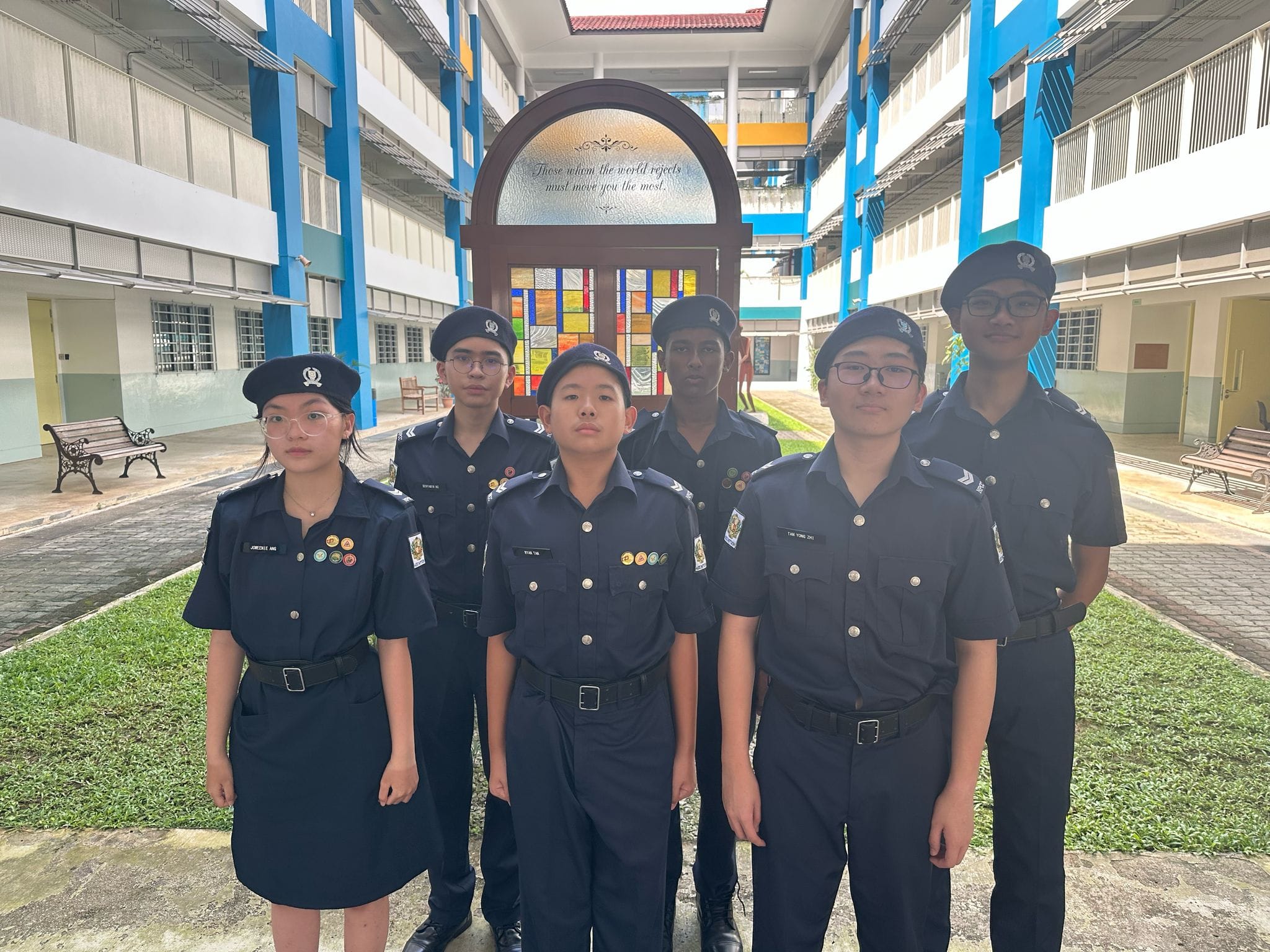 Six young students in dark navy uniforms and caps stand in a school courtyard in front of a decorative arched door.