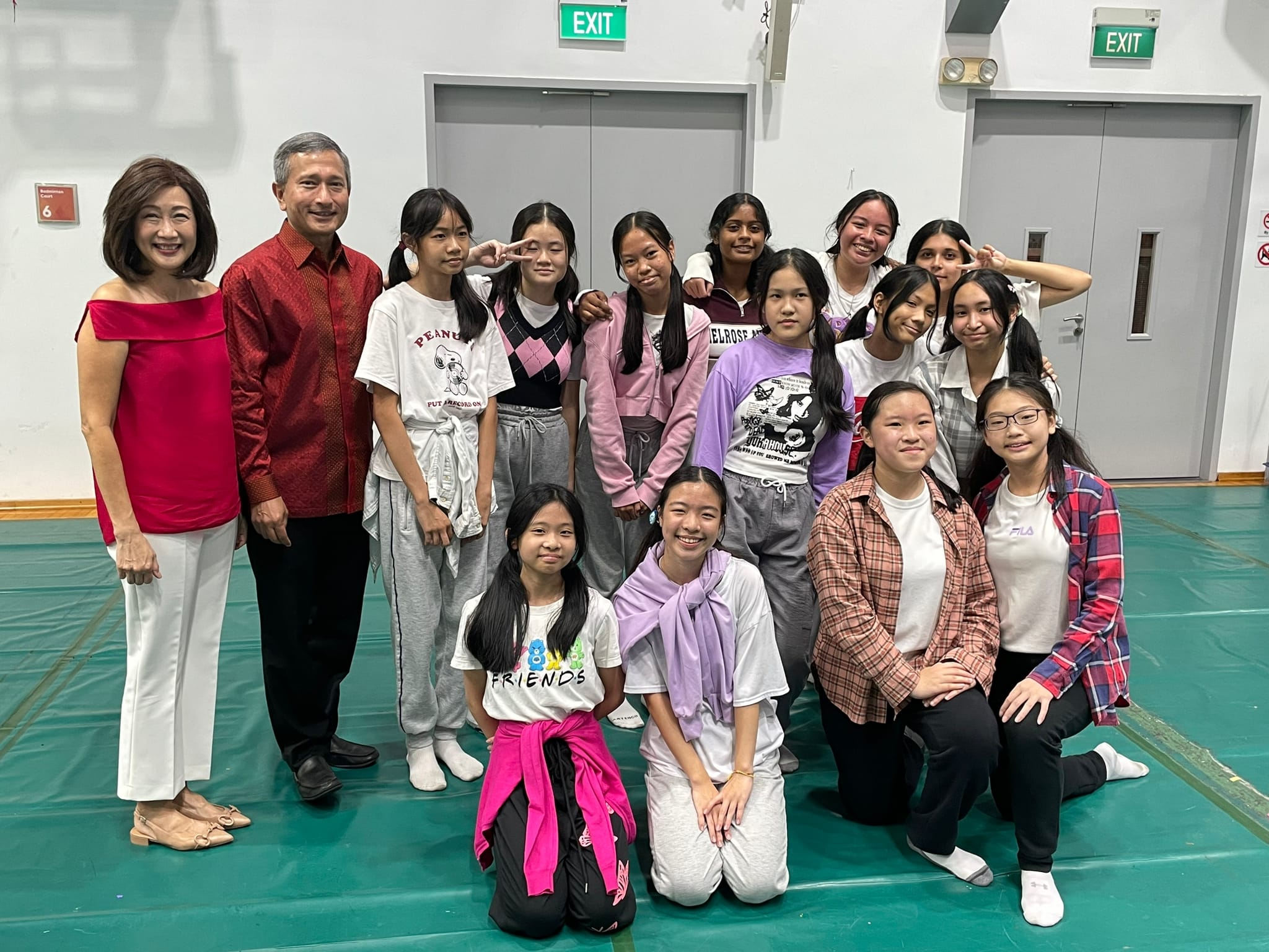 A man in red and a woman in red top pose with a group of teenage girls in a school gymnasium.