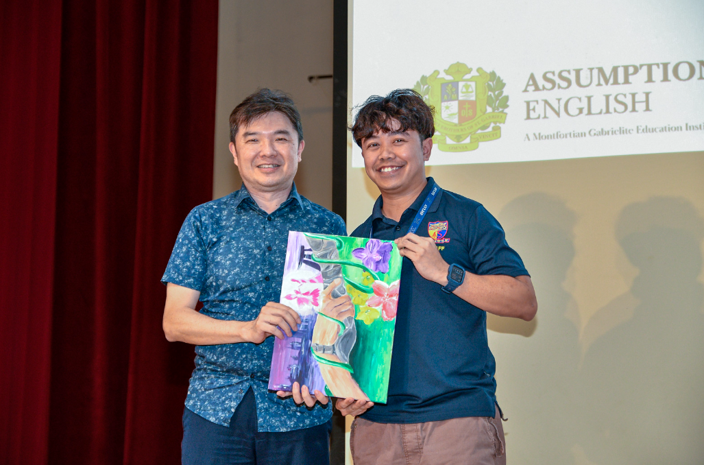 Two smiling men hold a colorful floral painting together in front of an Assumption English school banner.