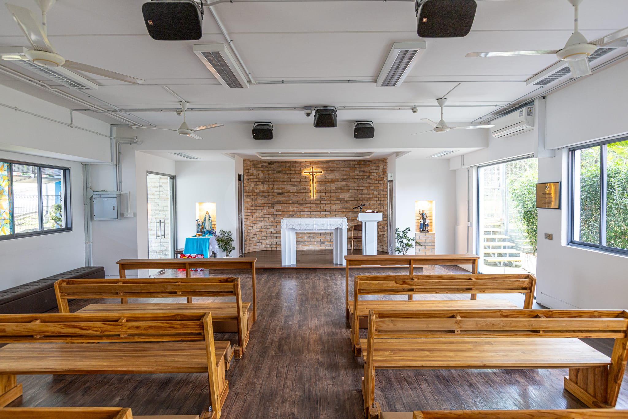 Small Catholic chapel interior with wooden pews, white altar, glowing crucifix on brick wall, and ceiling fans.