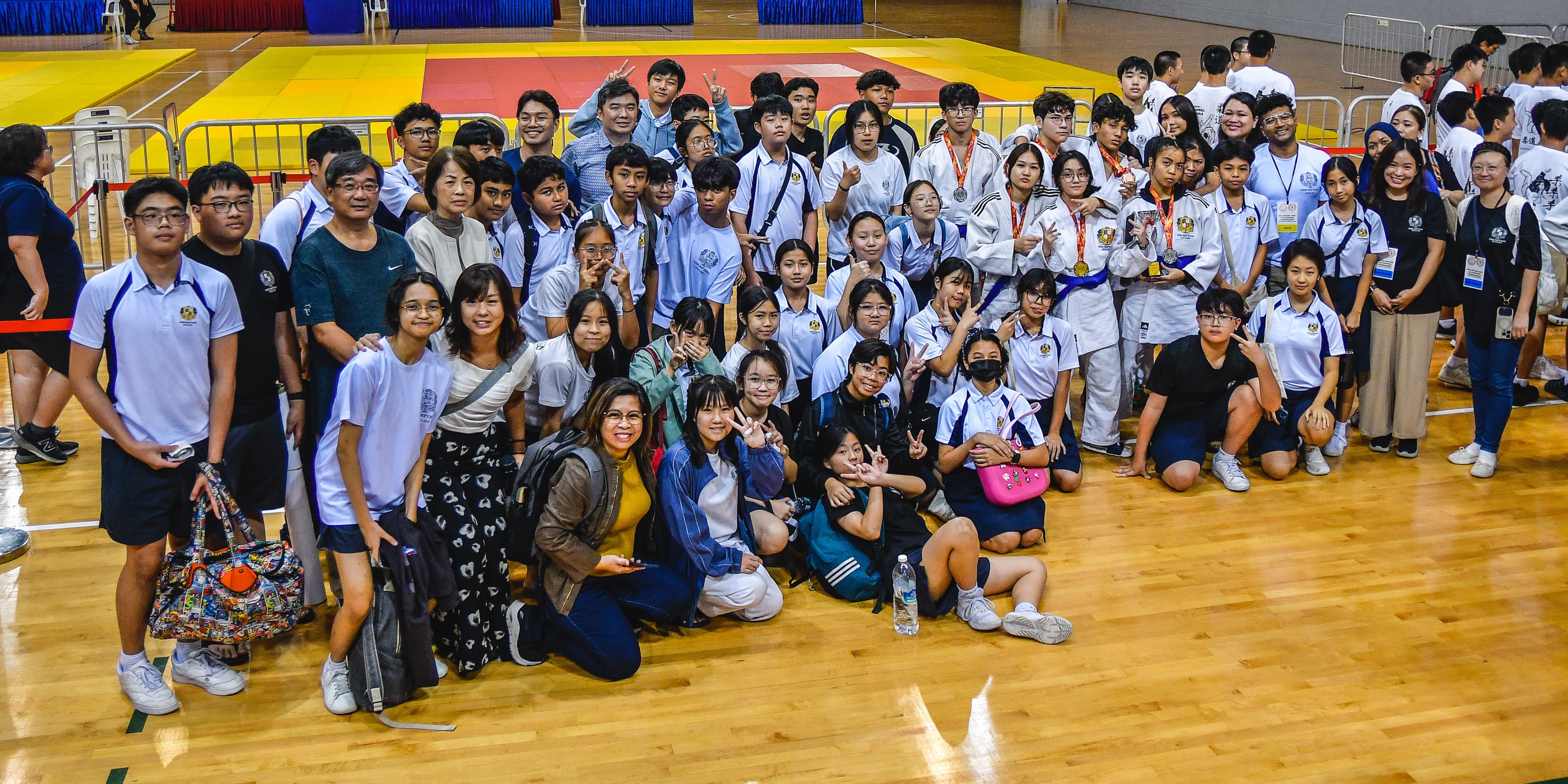 Large group of students and adults posing together in a sports hall with a colorful martial arts mat in the background.