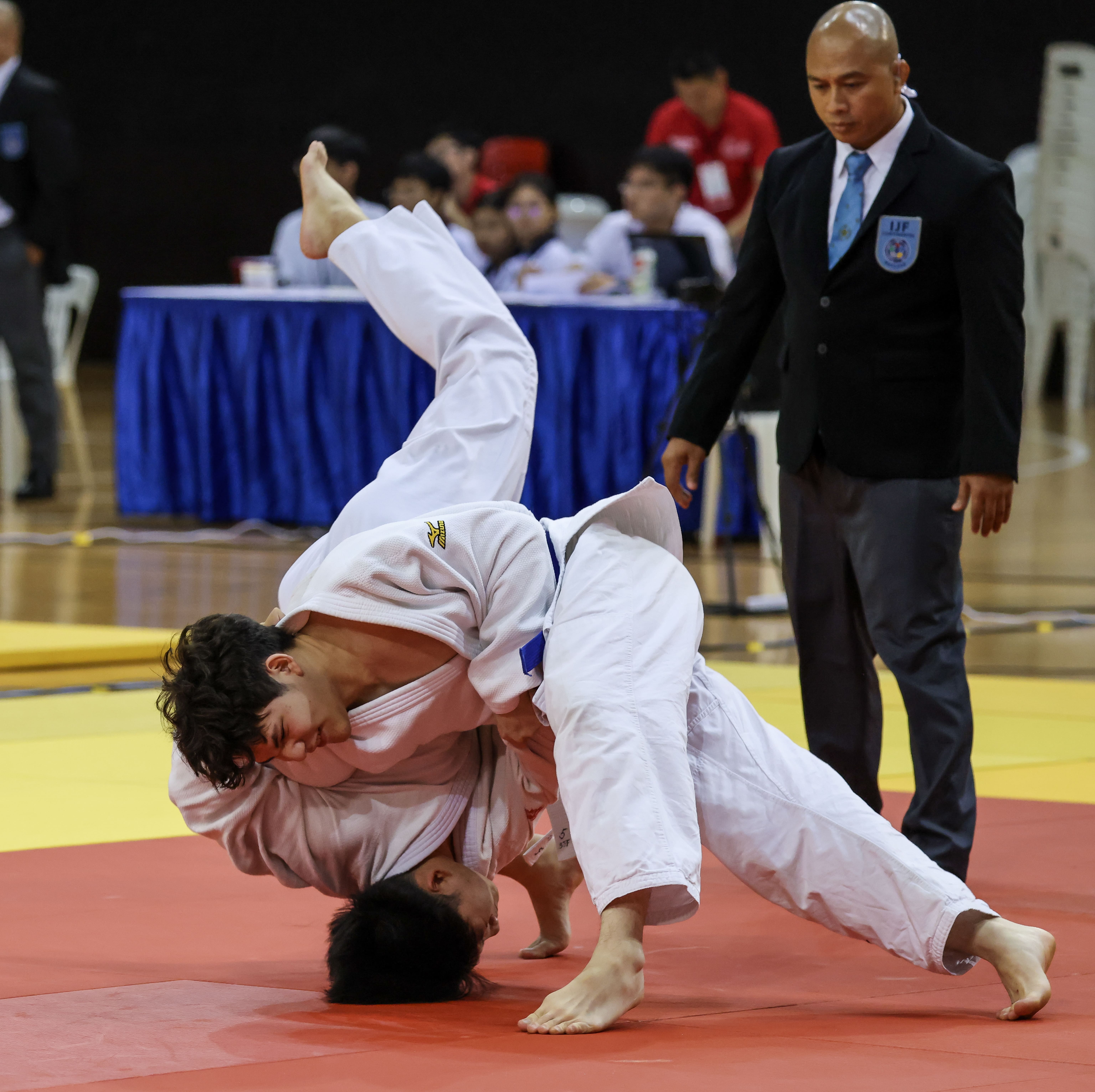 Two judo competitors in white uniforms grapple on a mat as a suited referee watches closely.