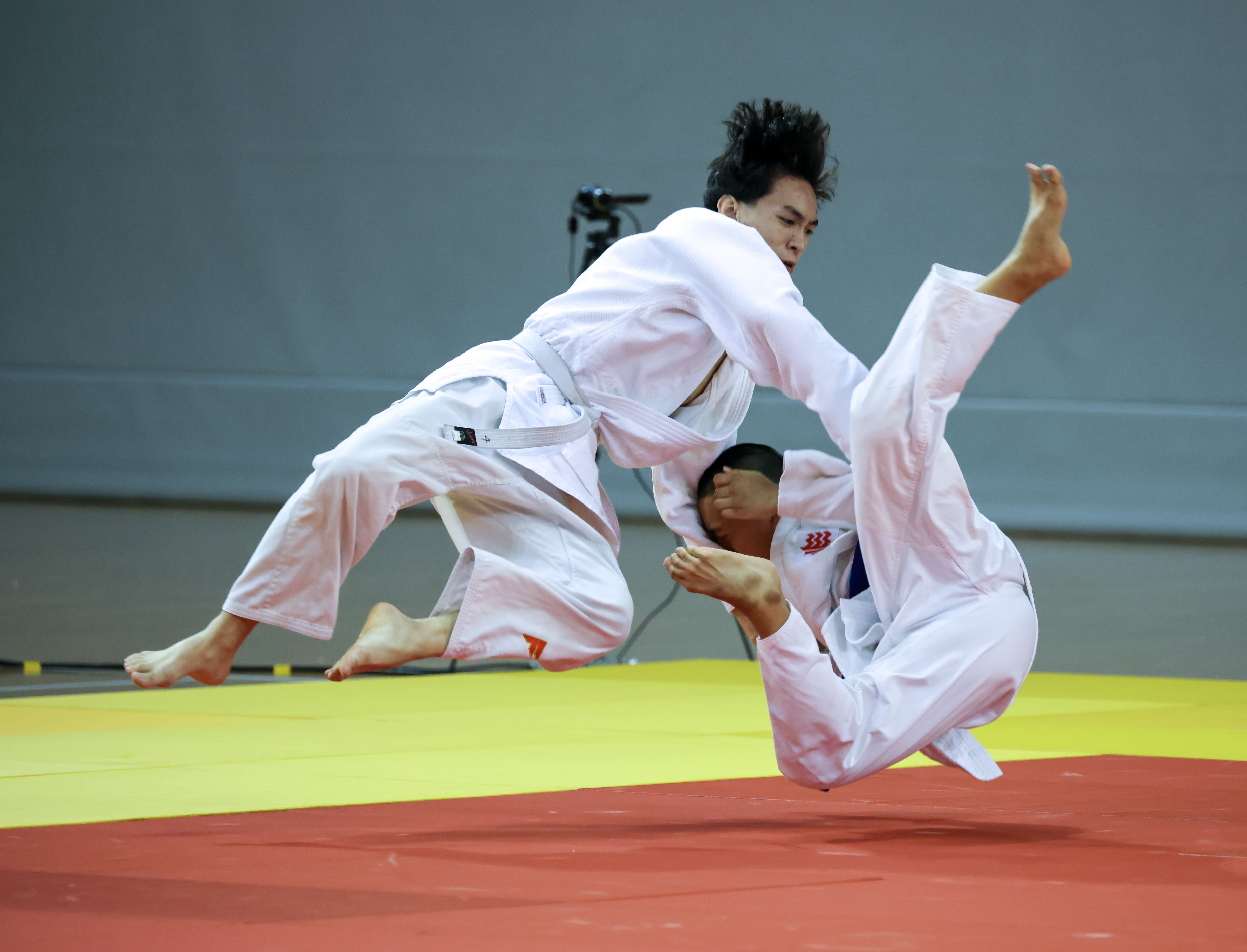 Two judo athletes in white uniforms mid-air as one throws the other onto a red and yellow mat.