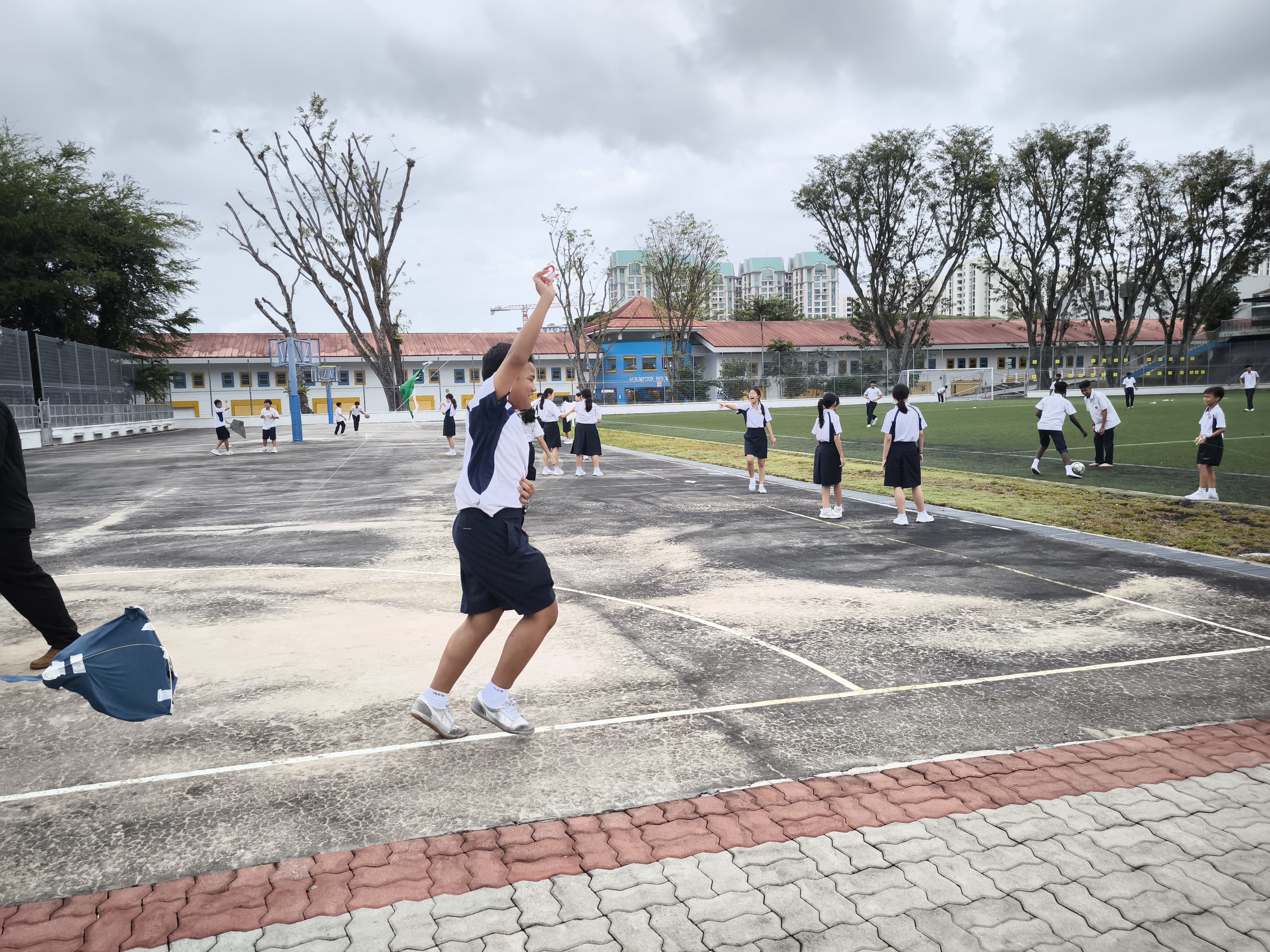 Students in white and navy uniforms play outdoors on a school court and grass field under a cloudy sky.