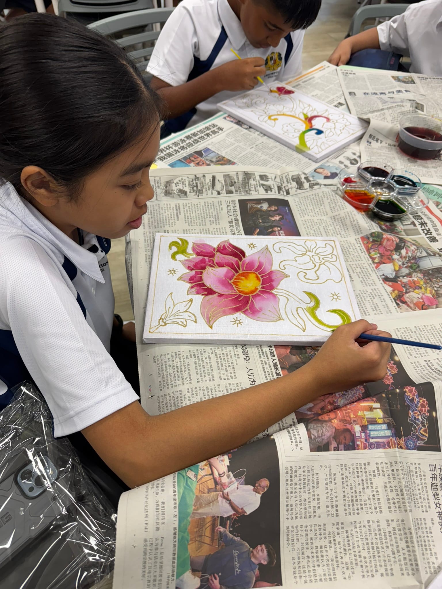 A young student in a white uniform paints a pink lotus flower on canvas over newspaper-covered tables.
