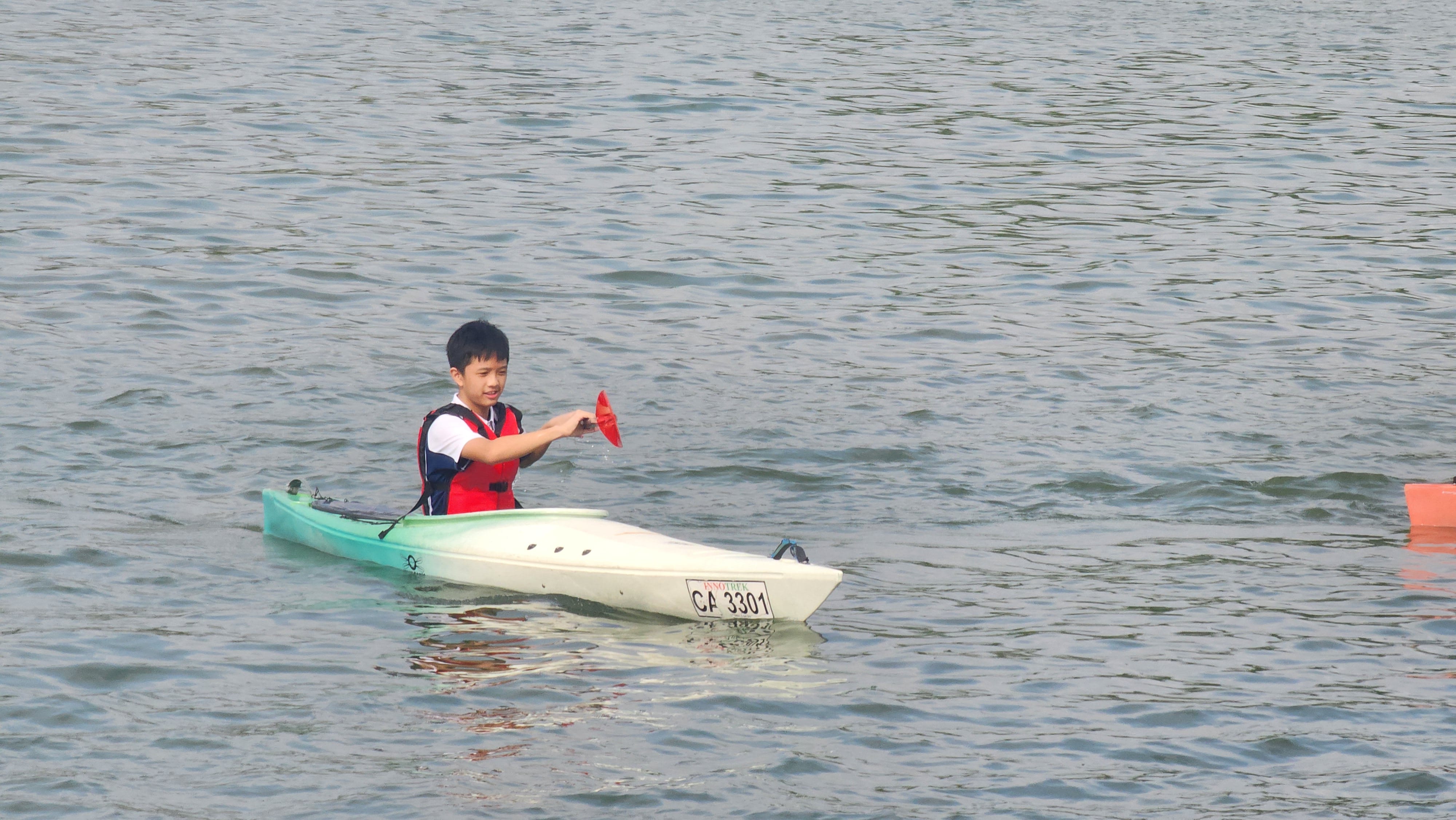 A young boy in a red life vest sits in a white and green kayak labeled CA 3301 on calm water.