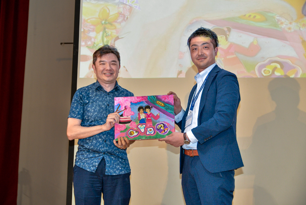 Two men smile while holding a colorful children's painting on a stage with a projected artwork background.