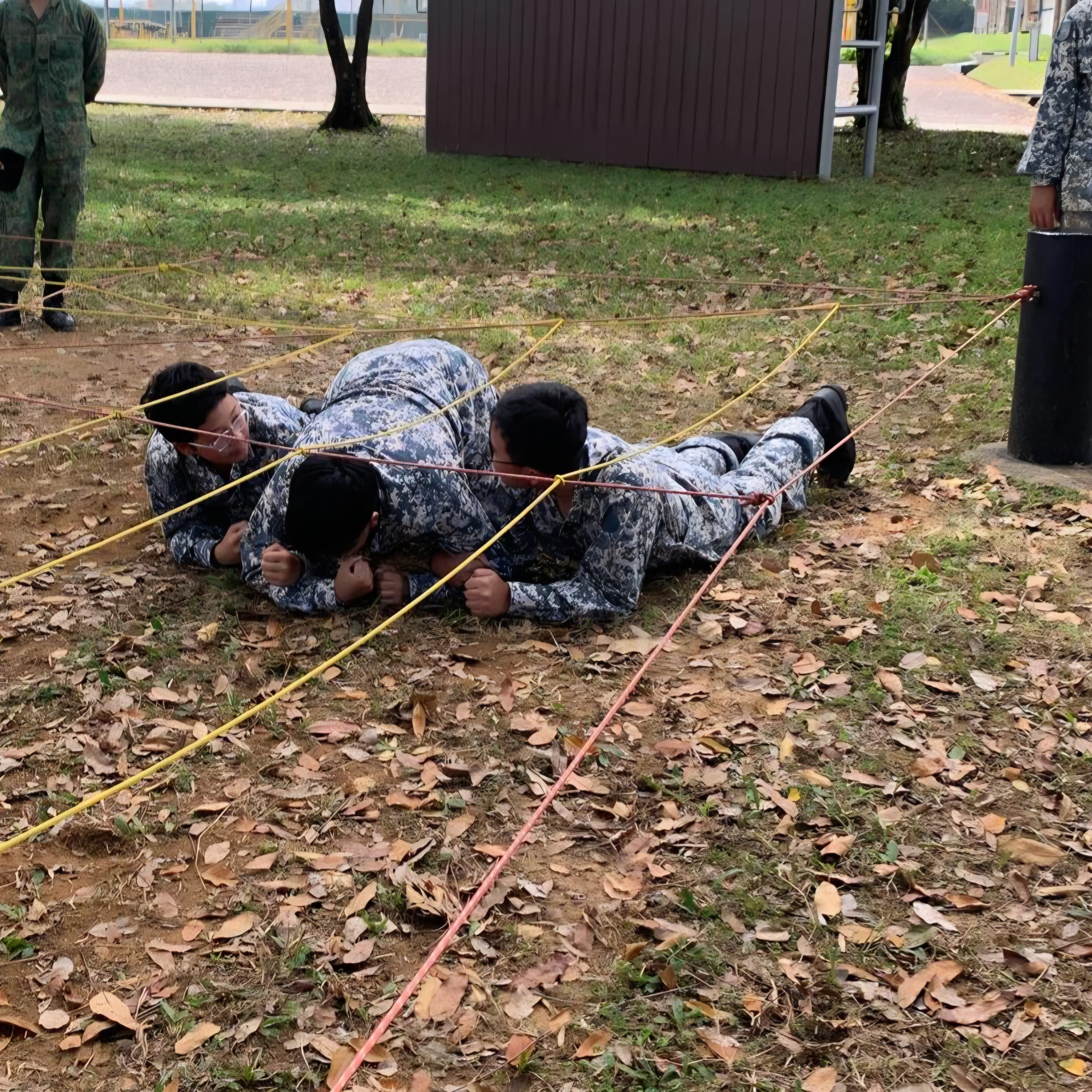 Three soldiers in camouflage uniforms crawl under low ropes on a leafy ground during an outdoor training exercise.