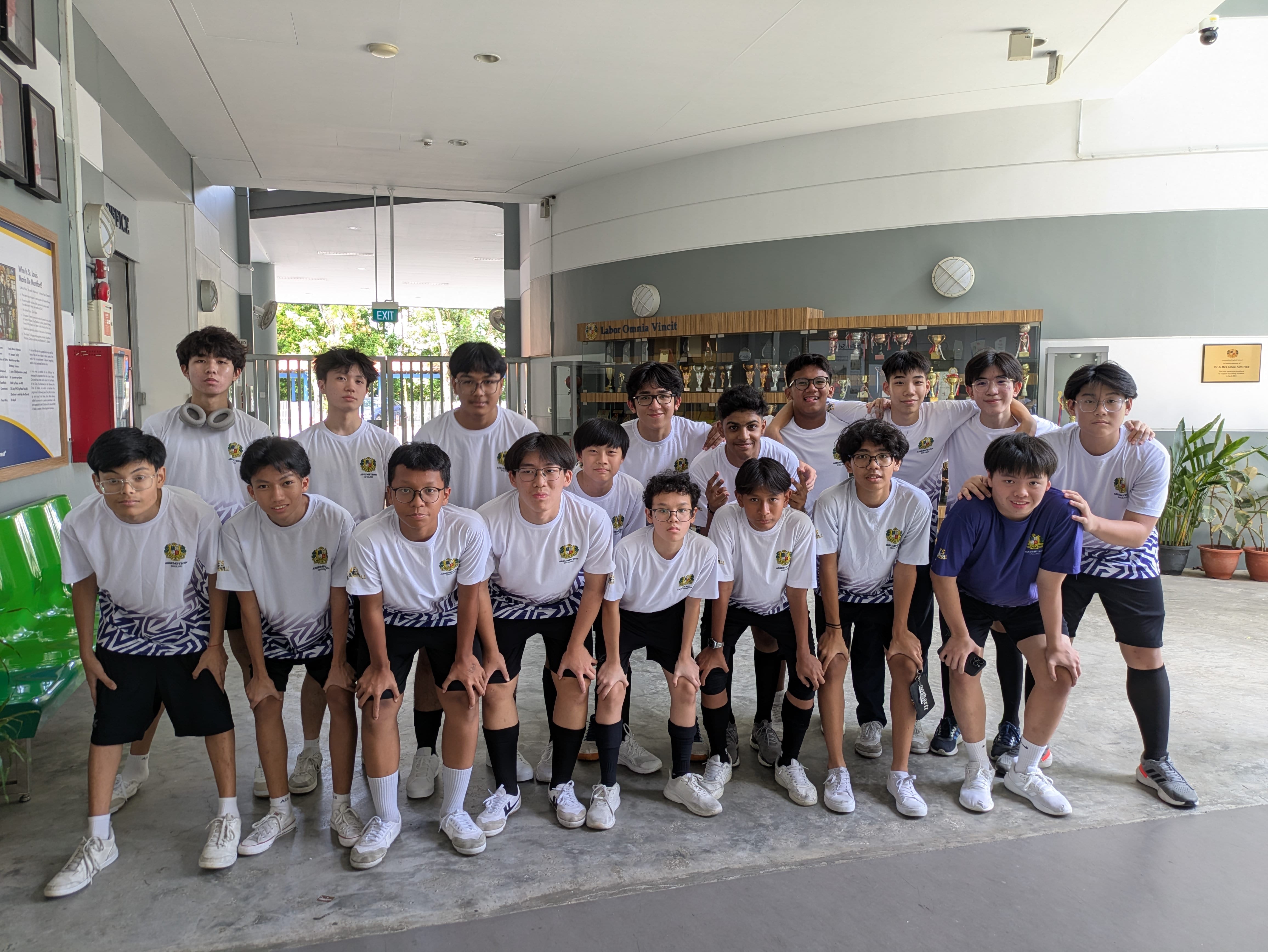 Group of teenage boys in white sports uniforms posing in two rows inside a school building with trophy cabinets behind them.