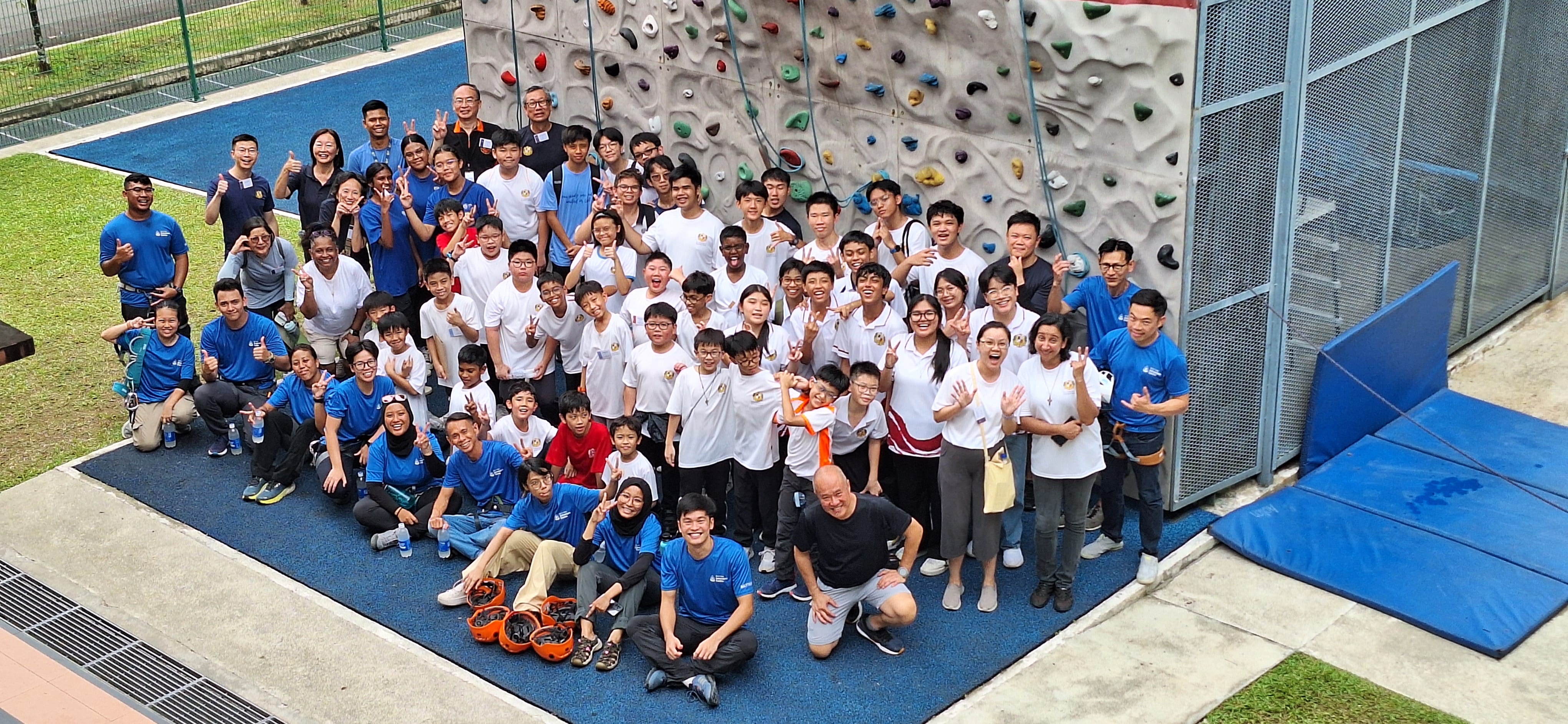 Large group of children and adults posing in front of an outdoor rock climbing wall, many making peace signs.