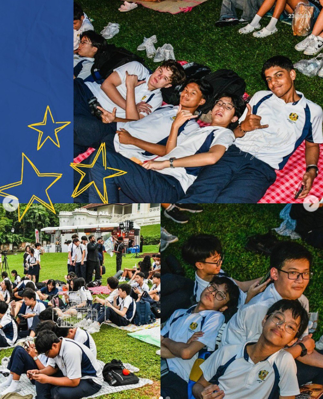 Students in white and navy uniforms relax on grass during an outdoor school event with a stage in the background.