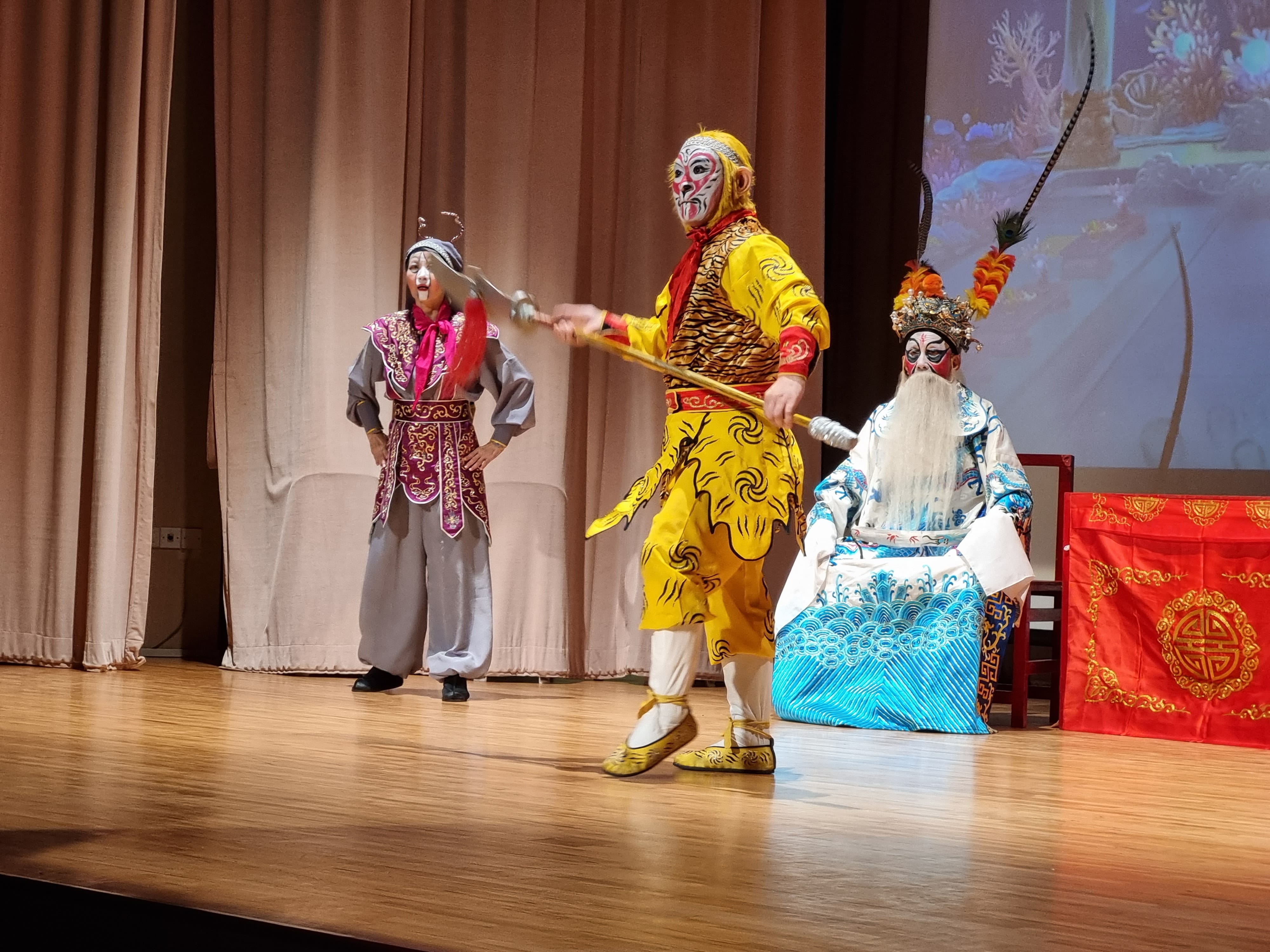 Three performers in colorful Chinese opera costumes and face paint perform on a wooden stage.