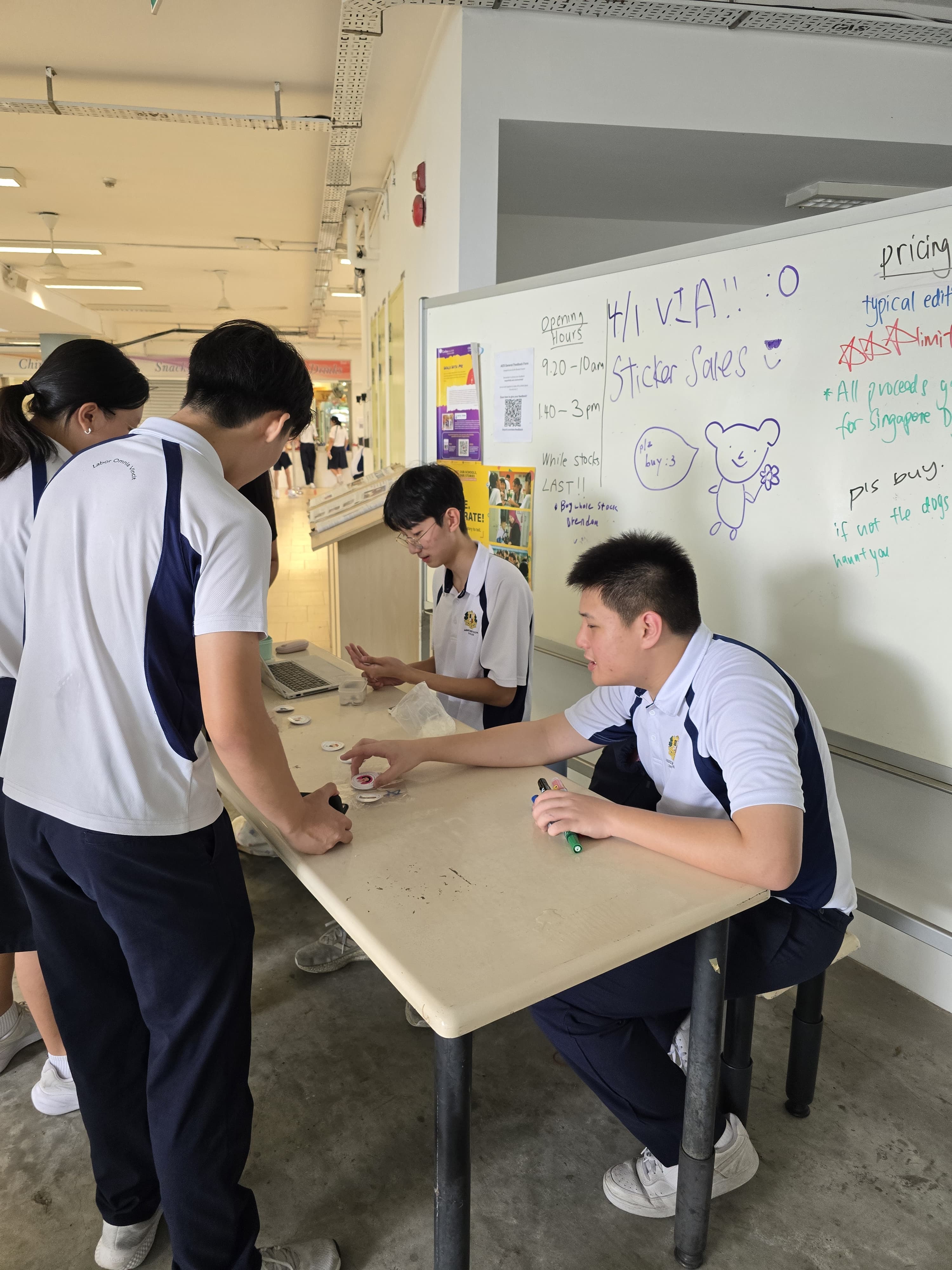 Students in school uniforms run a sticker sales booth at a table with a whiteboard showing pricing and hours.