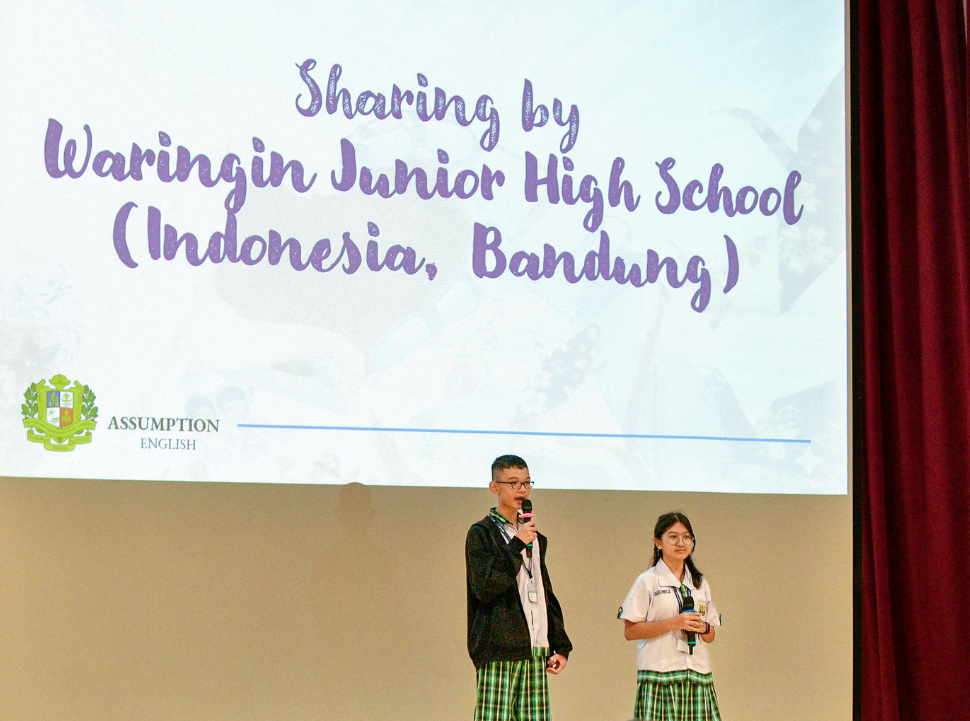 Two uniformed students present on stage with a slide reading "Sharing by Waringin Junior High School (Indonesia, Bandung)."