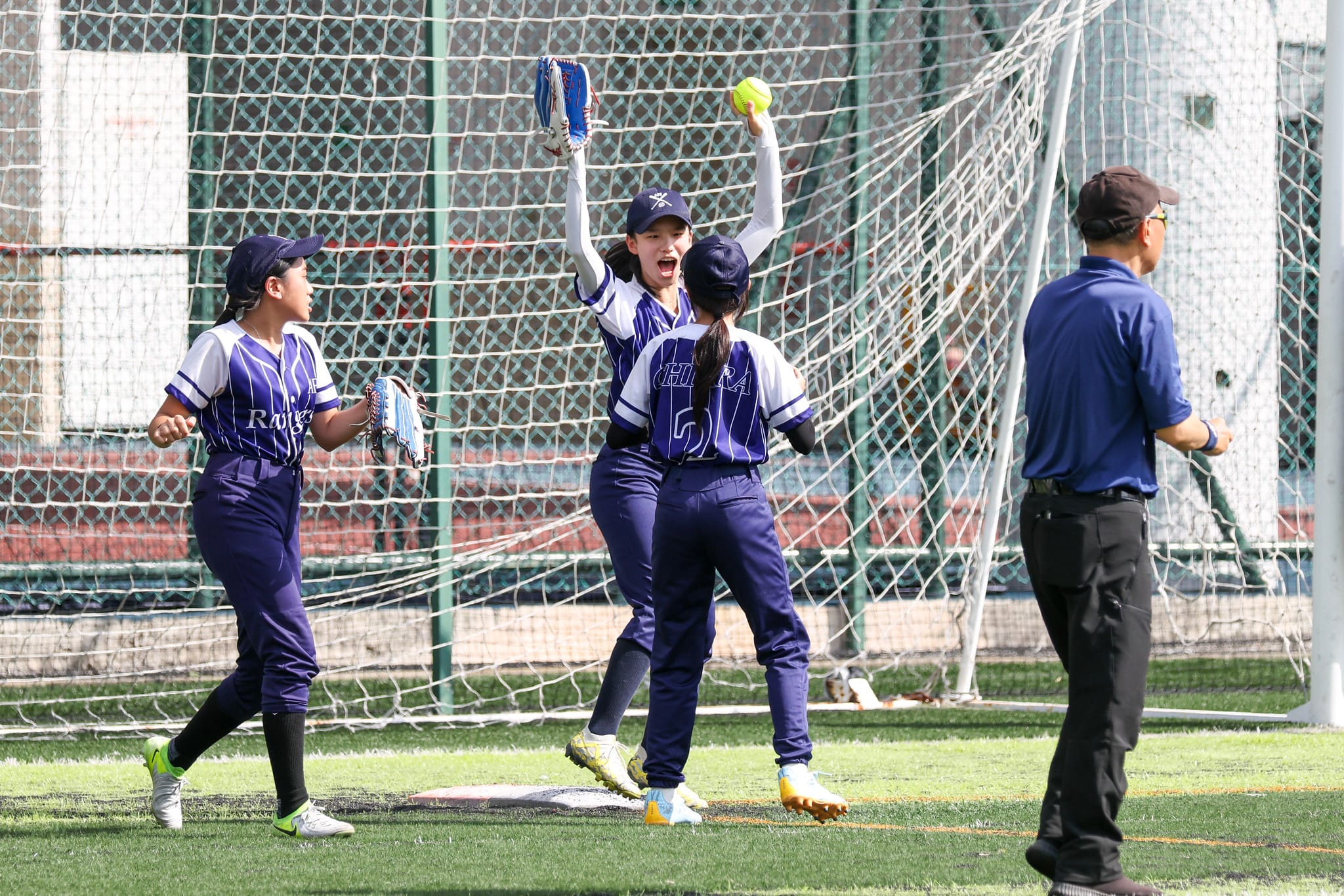 Three female softball players in blue uniforms celebrate on a field, one raising a yellow ball, as an umpire watches.
