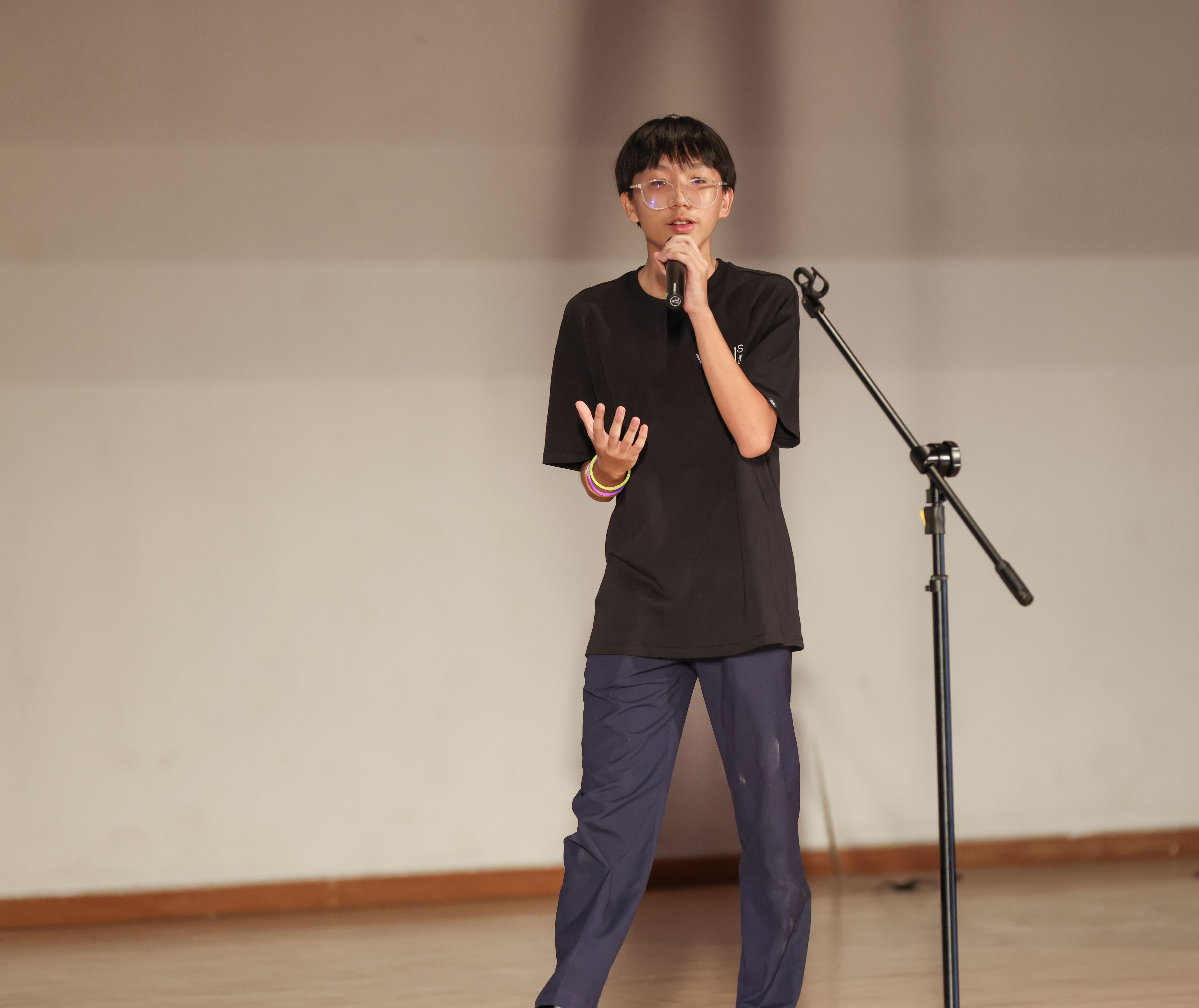 A young person wearing glasses and a black shirt speaks into a handheld microphone on a stage near a mic stand.