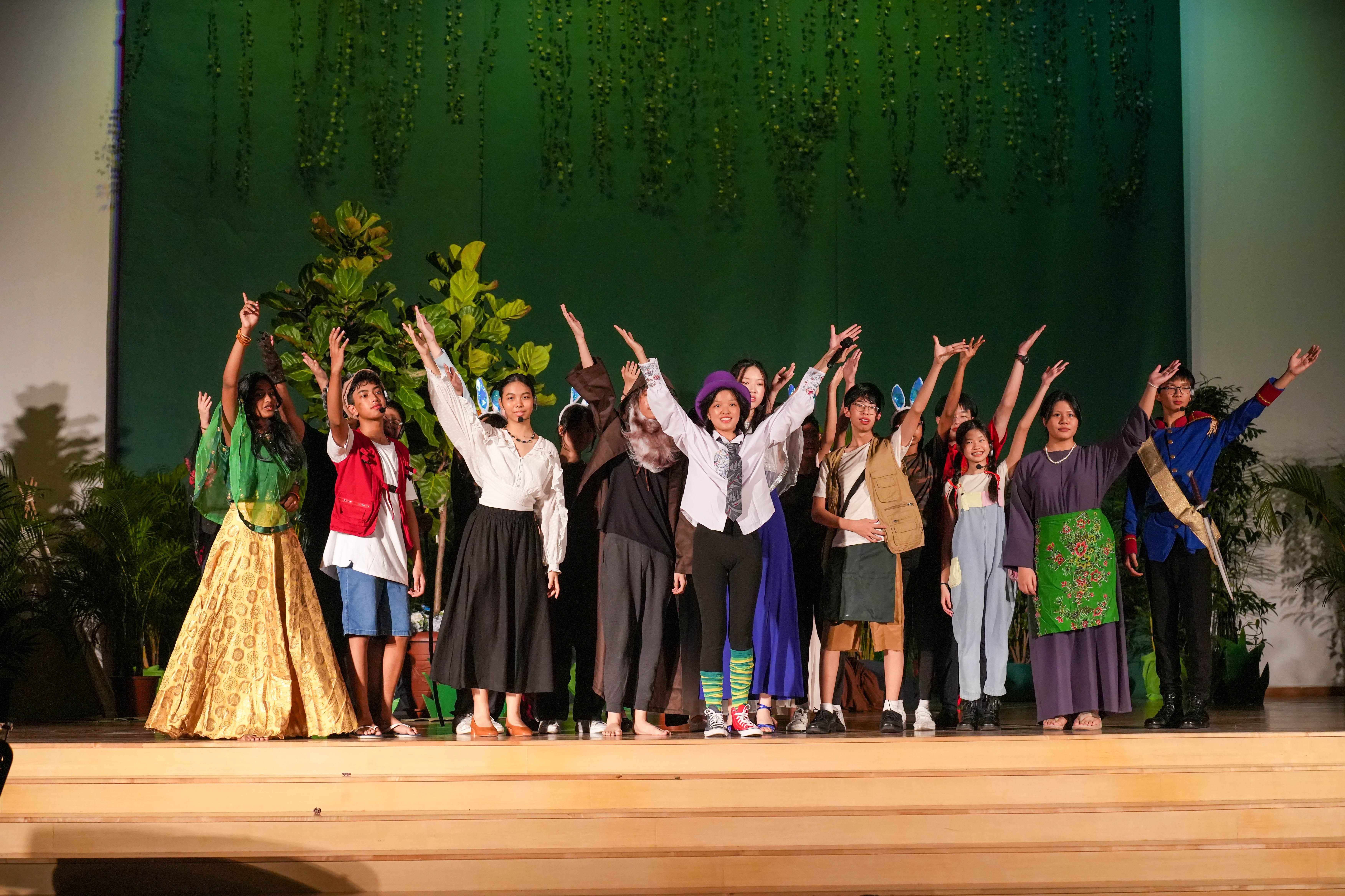 A group of young performers in colorful costumes raise their arms on a stage with a green leafy backdrop.