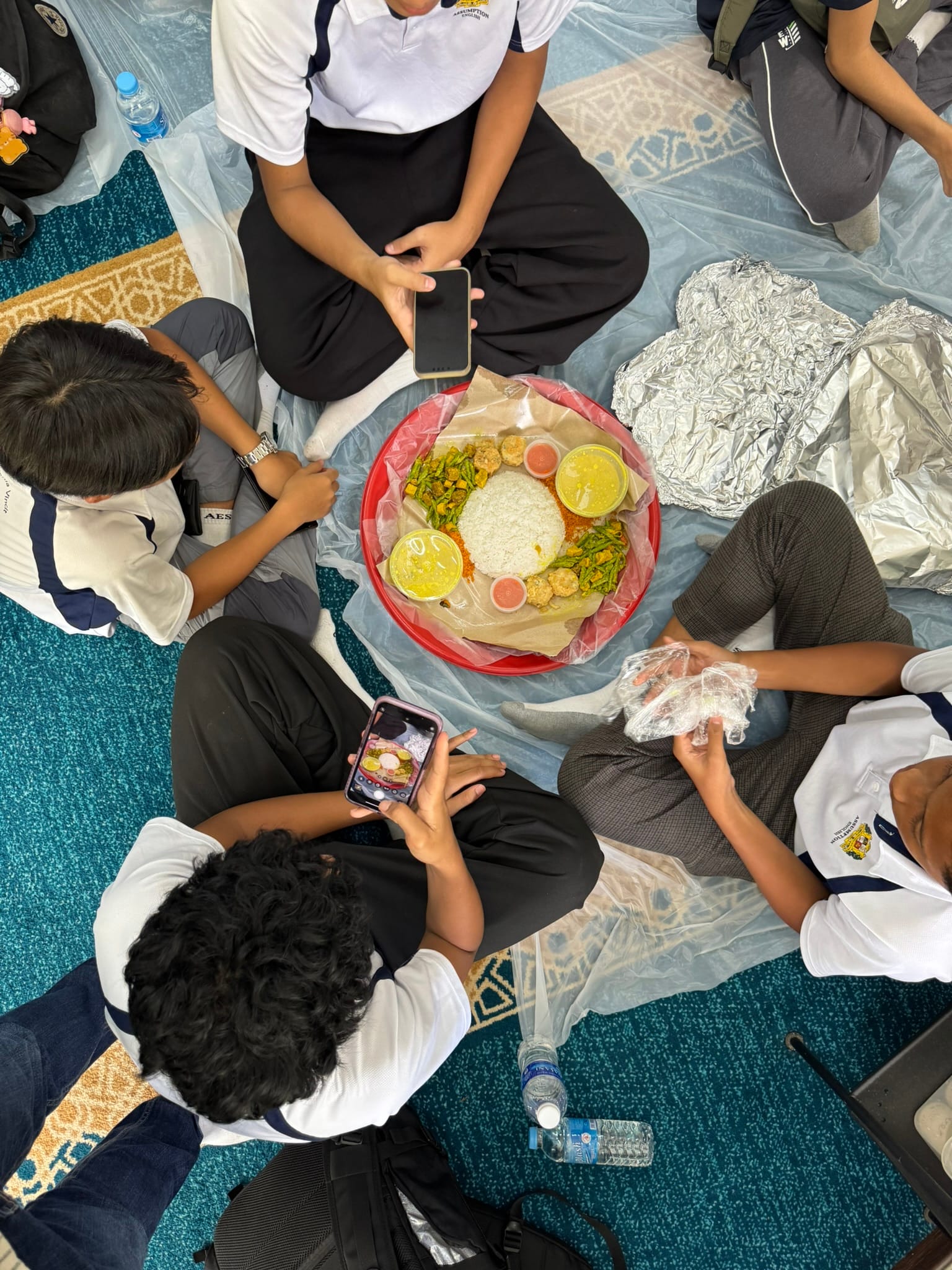 Students in school uniforms sit on the floor around a large shared plate of rice and vegetables, with one photographing the food.