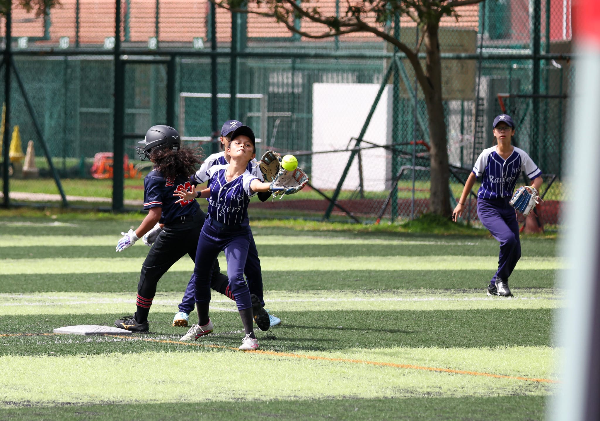 A youth softball fielder in a Rangers uniform catches a yellow ball near a base as a runner arrives.
